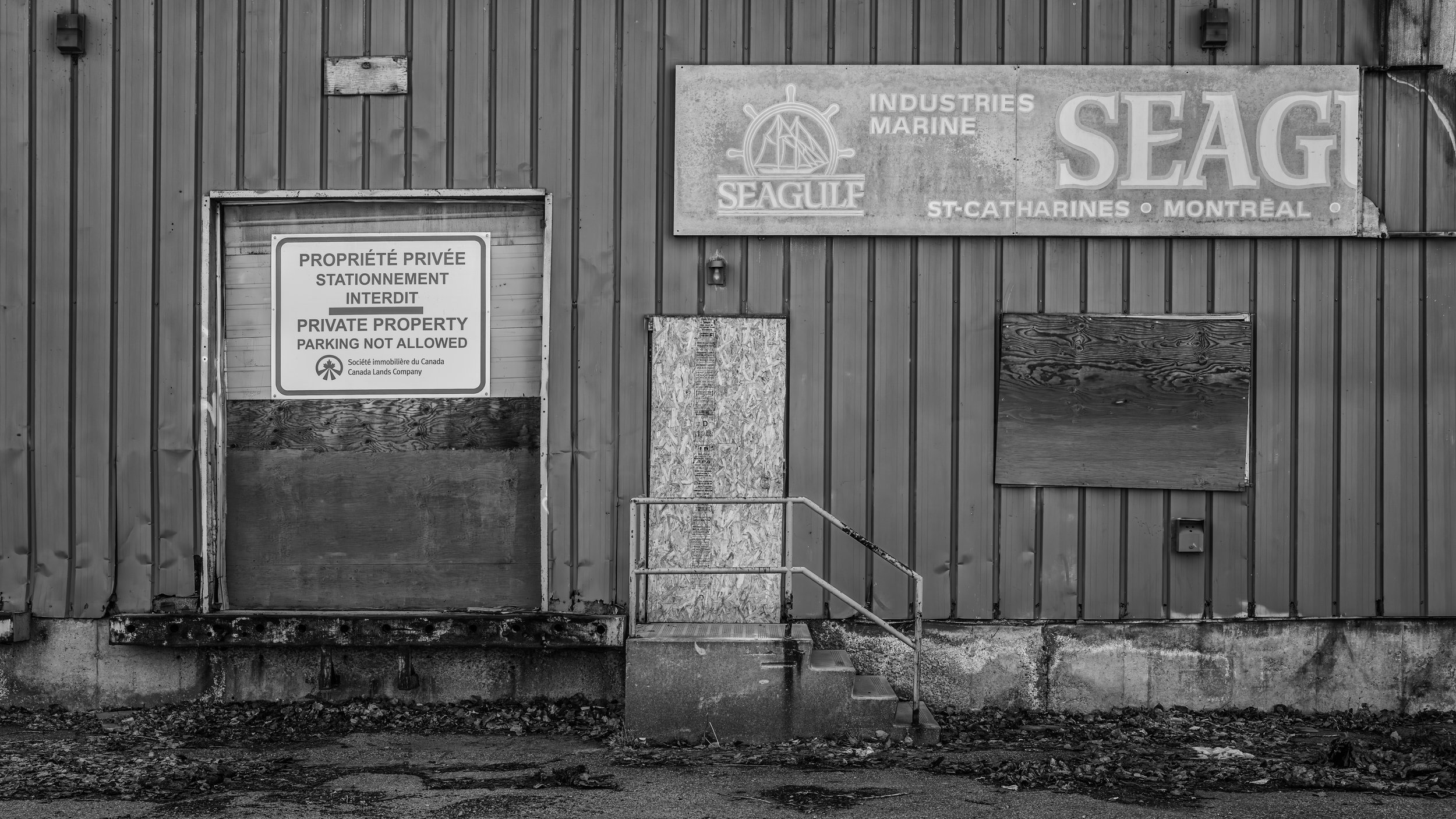 A black and white photo of a building with wood and metal siding, featuring a door with a sign that says 'Private Property Parking Not Allowed' in French and English. The door appears boarded up with wood panels, and there are a few steps leading up 