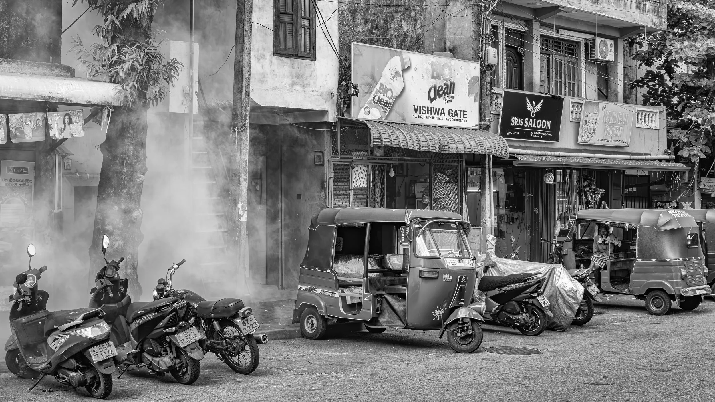 Street scene with parked motorcycles and auto rickshaws, buildings with signs including 'BIO clean', 'VISHWA GATE', and 'SRIS SALOON', a large tree, and some smoke or fog in the air.