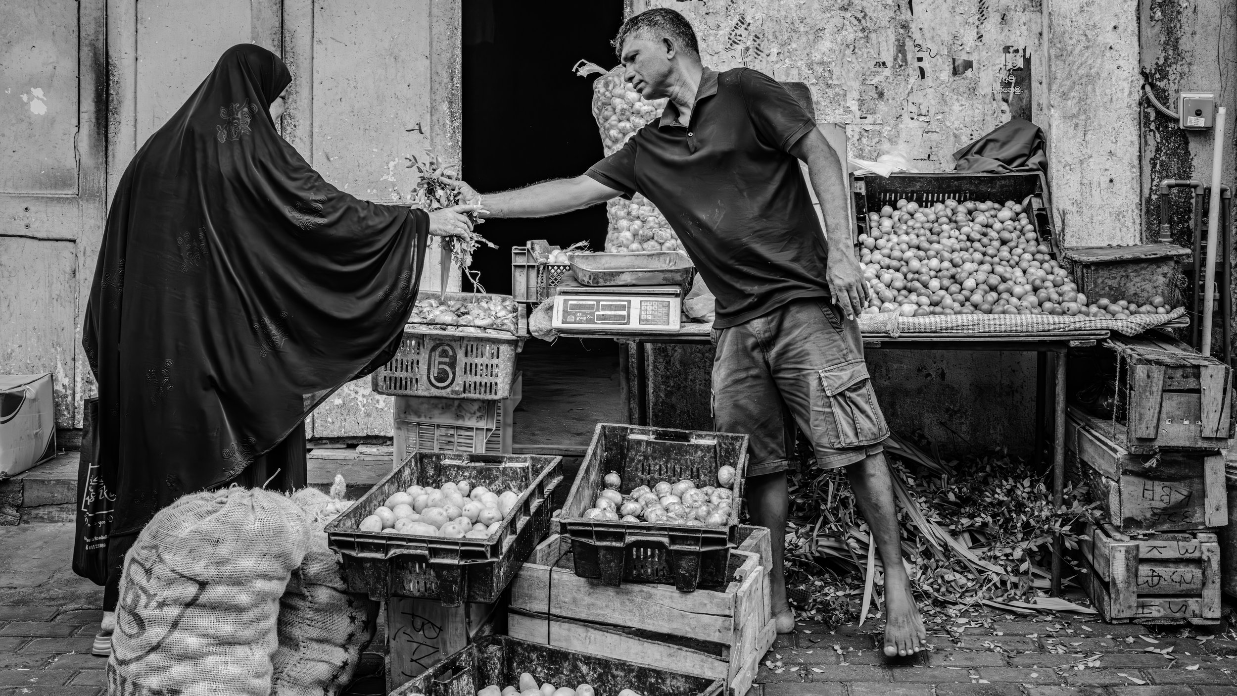 A street market scene in black and white, showing a man handing a bunch of vegetables to a woman dressed in a long, dark, embroidered dress and a headscarf. Colombo, Sri Lanka.