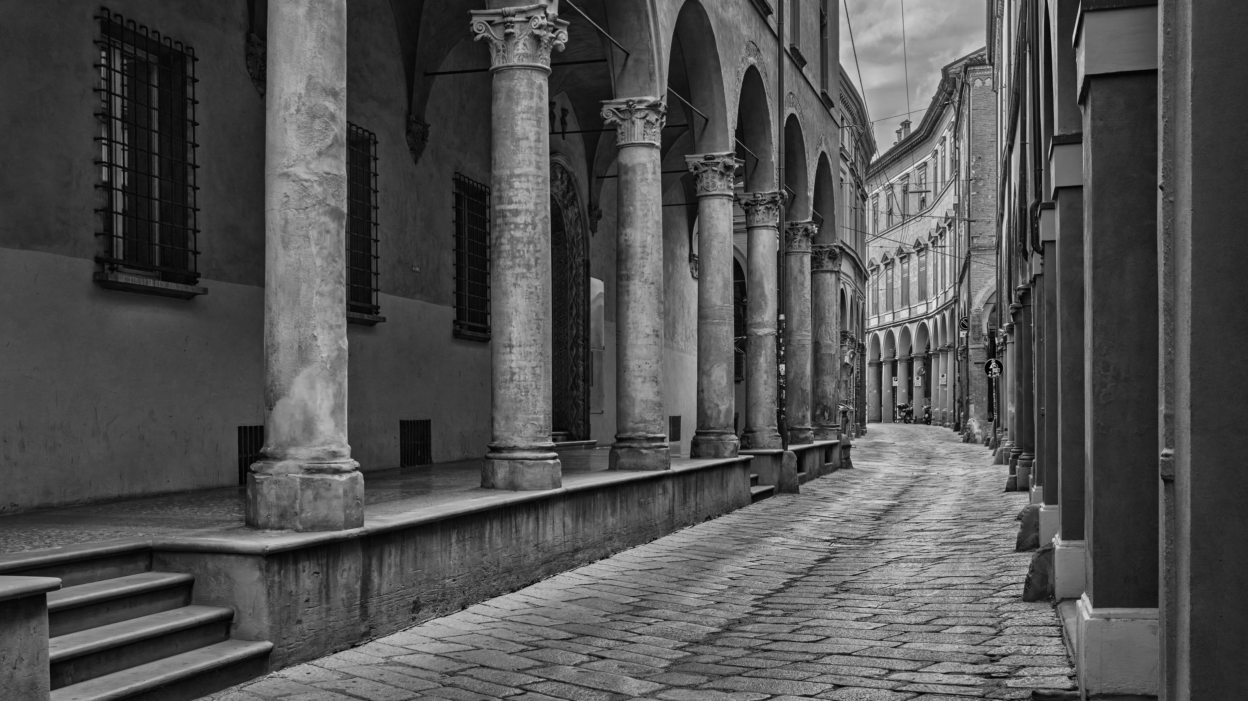 A narrow, quiet street lined with historic buildings and stone columns, with a cobblestone surface and window bars, under a cloudy sky. Bologna, Italy.