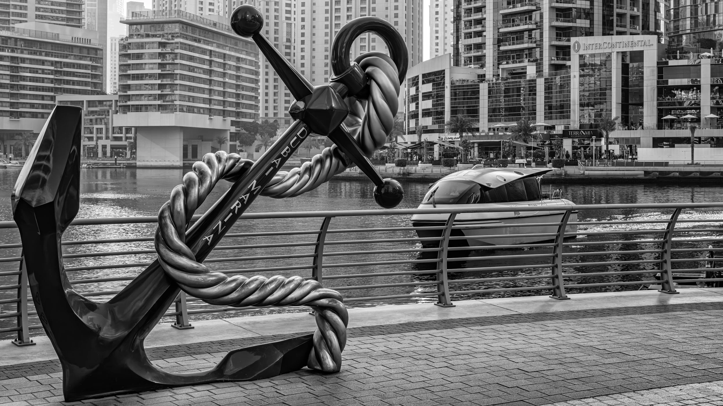 A large nautical-themed anchor sculpture with a twisted rope is positioned on a waterfront promenade, with modern high-rise buildings, a boat on the water, and a railing in the background.