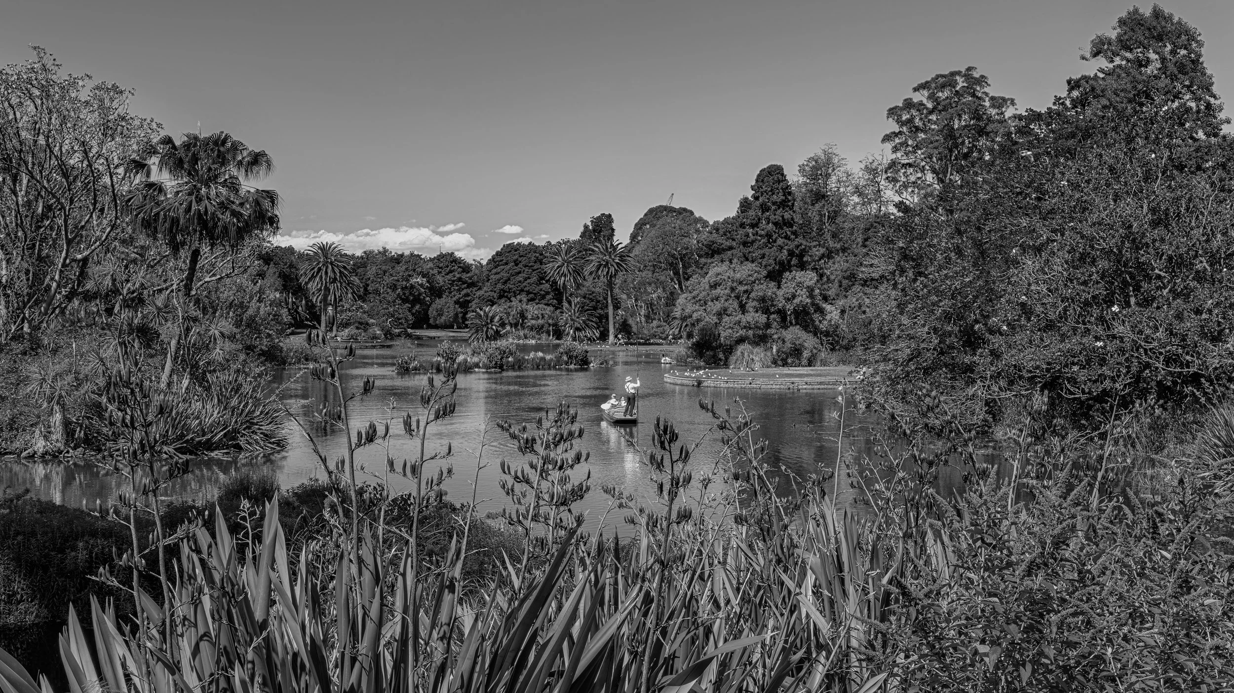 A black and white photograph of a tranquil river surrounded by lush trees and plants. Two people are in a small boat on the river, and a woman stands in the boat holding an umbrella. Tall grass and shrubs grow along the riverbank, and the sky is part