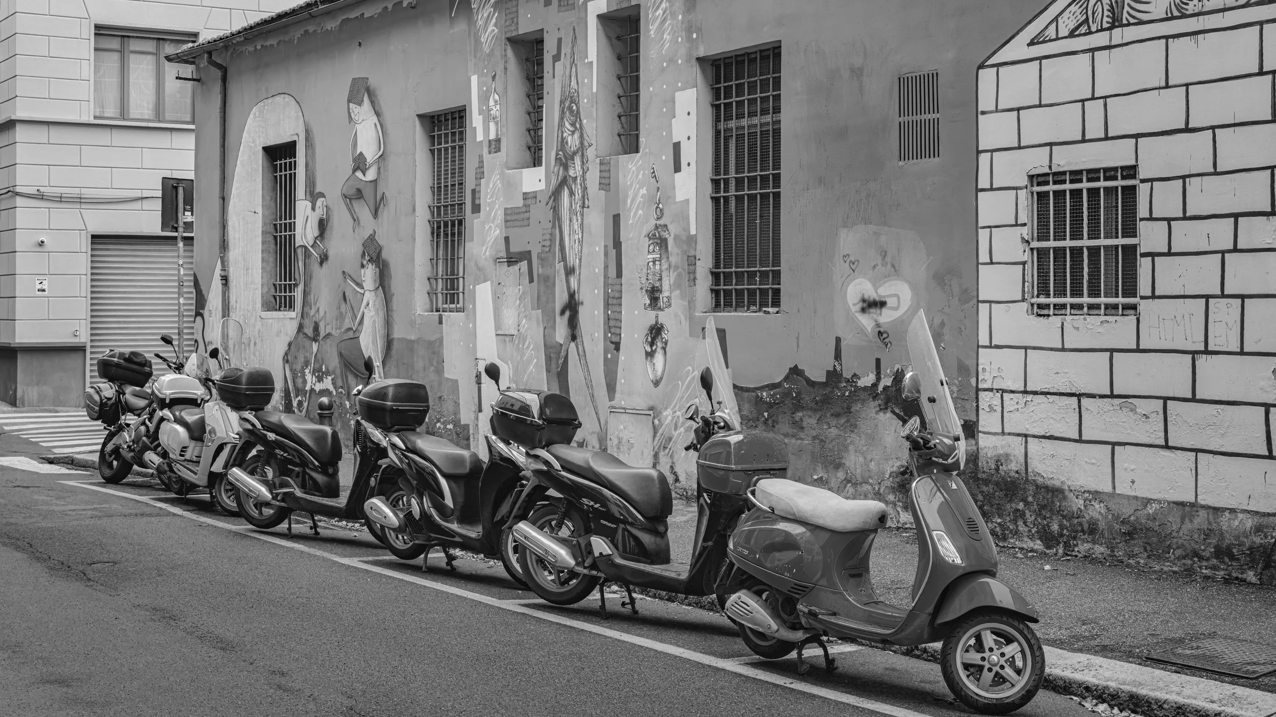 A row of parked scooters along a city street with a graffiti-covered wall in the background featuring various figures and symbols in black and white.