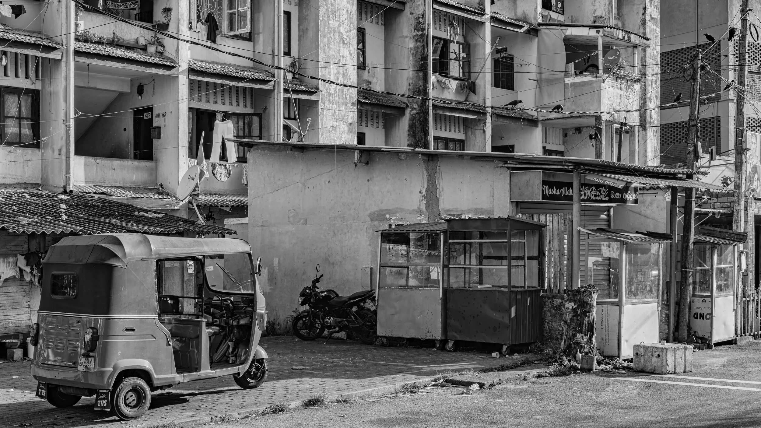 A black and white photo of a residential area showing an apartment building with balconies, some with laundry hanging and potted plants, a parked motorbike and an auto rickshaw in front of the building, a small structure that looks like a bus stop sh