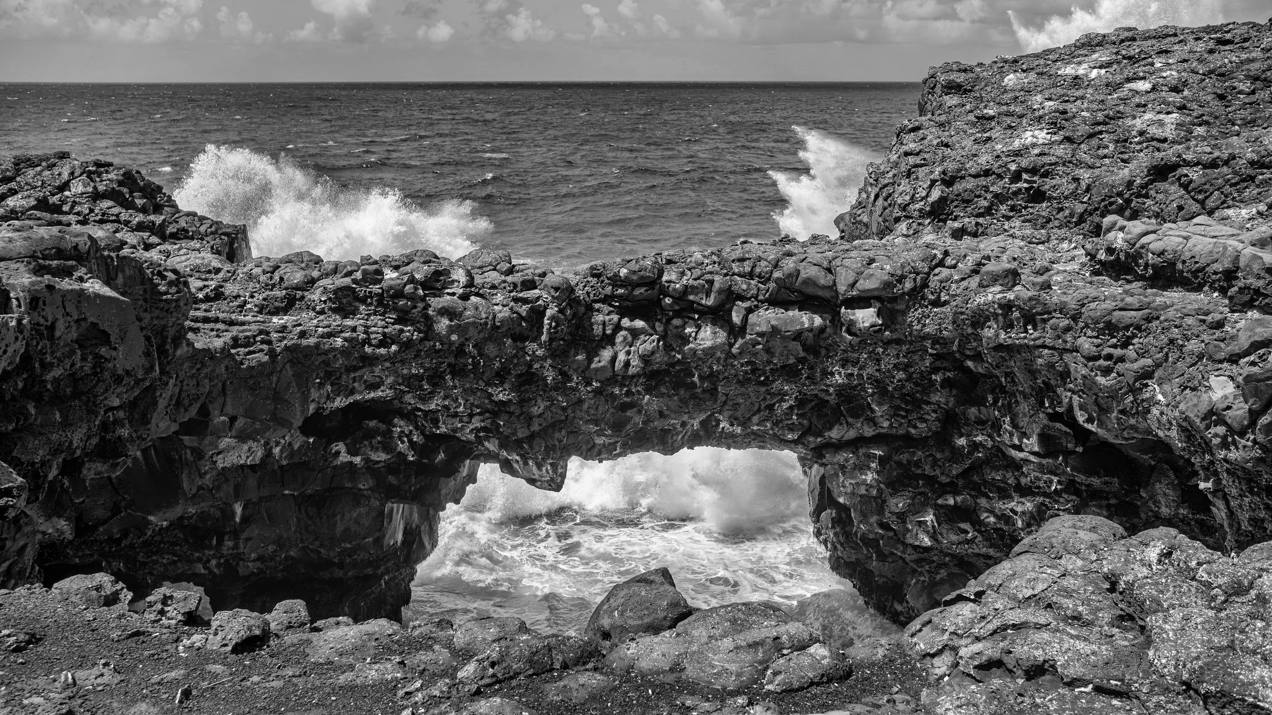Black and white photo of rocky coastal landscape with ocean waves crashing against rocks and a small arch formation in the foreground under a cloudy sky. Pont Du Naturel, Mauritius.