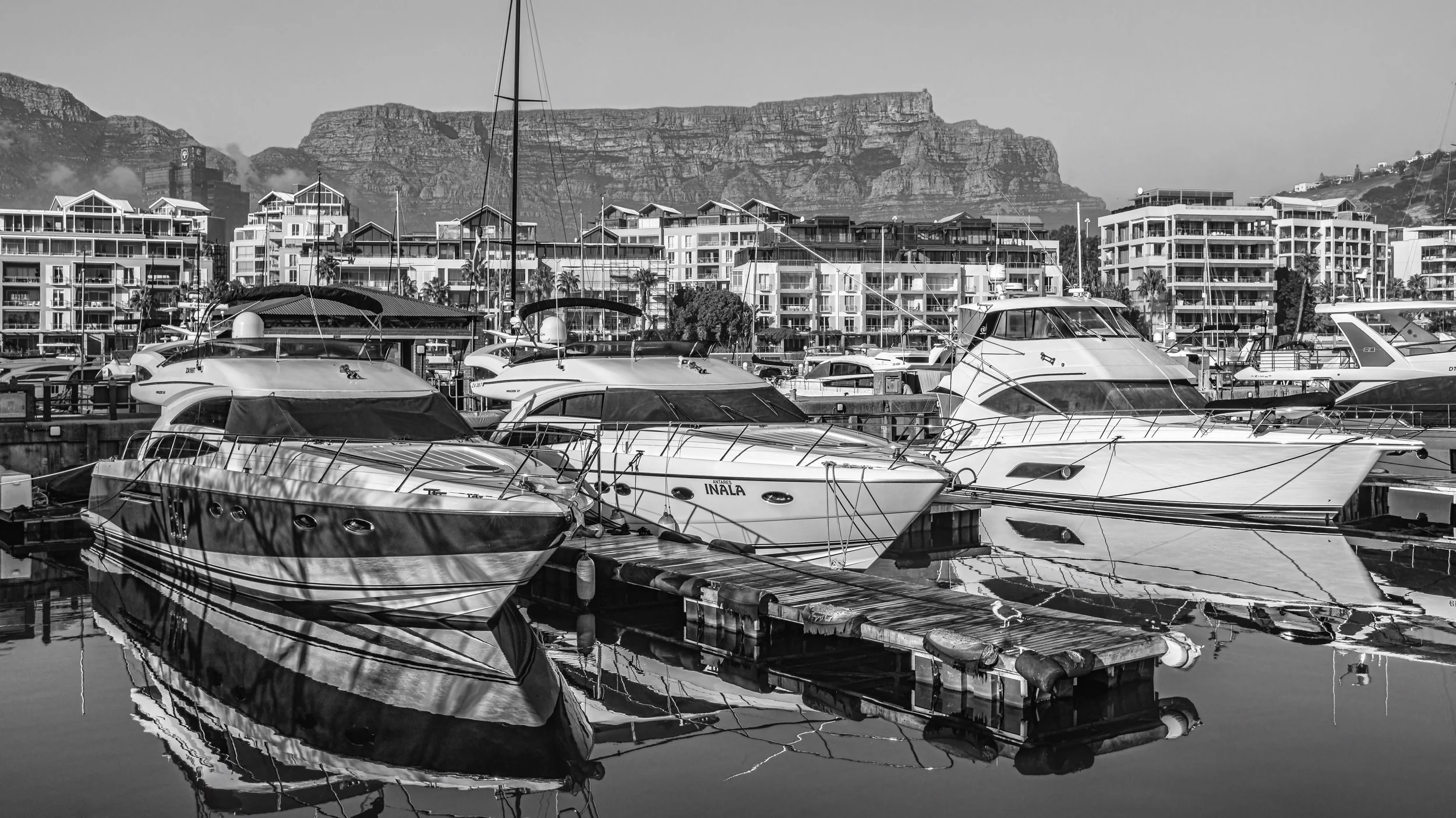 Black and white photo of boats docked at a marina with modern apartment buildings in the background and mountains in the distance. Cape Town, South Africa.