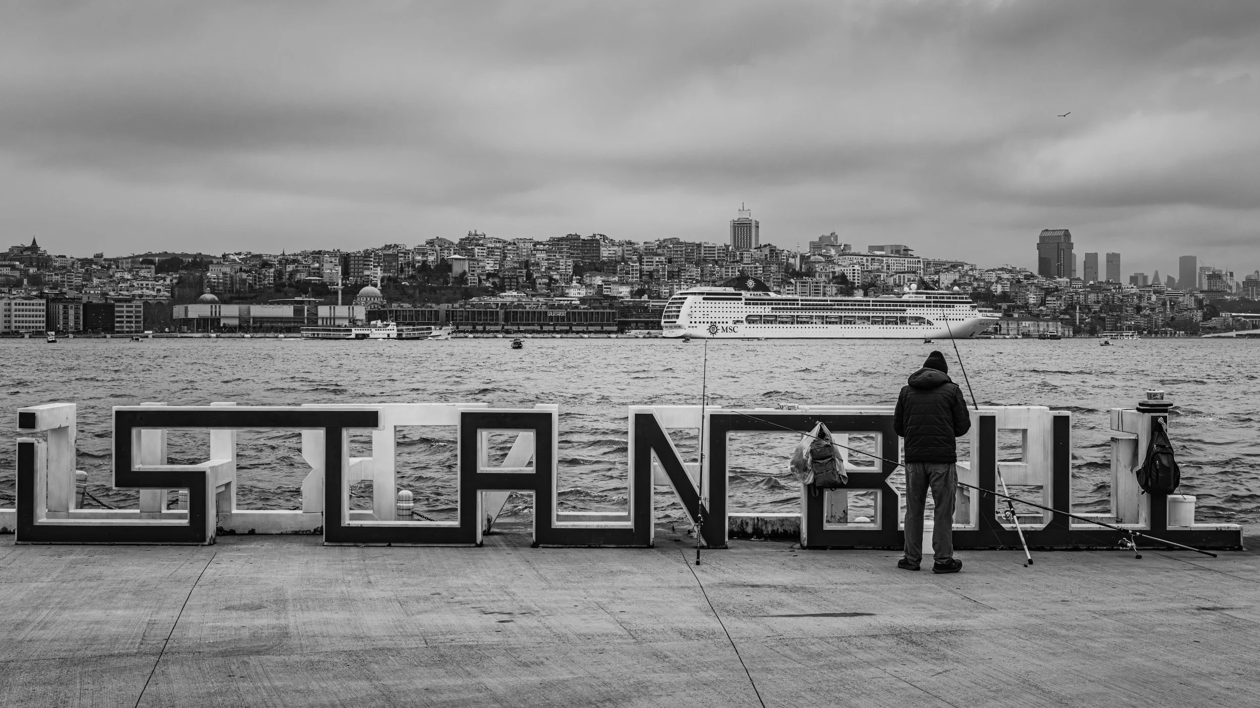 A person fishing on a concrete waterfront with large city buildings and a cruise ship in the background, under a cloudy sky, in black and white.