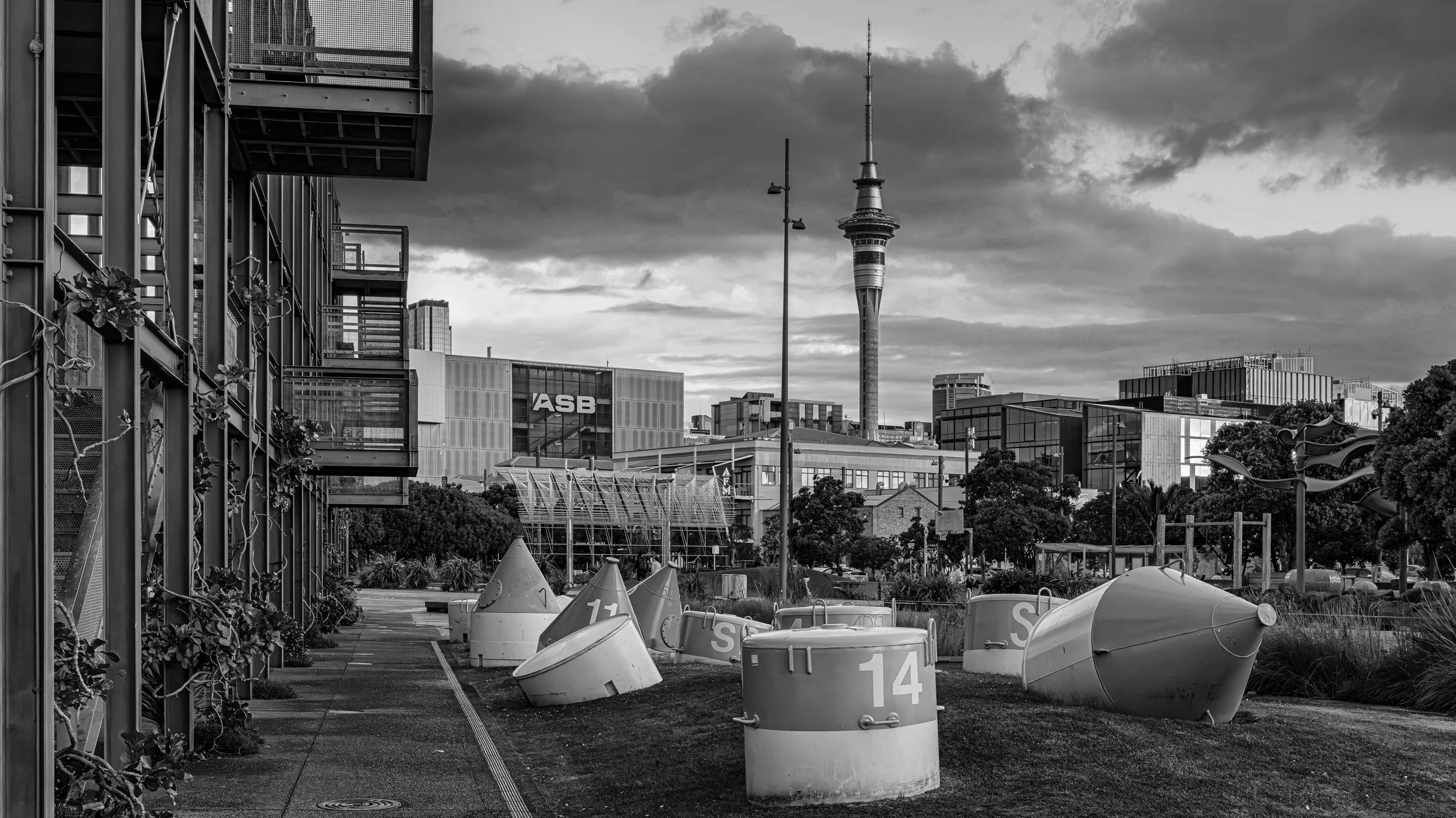 Cityscape with modern buildings, a tower with a spire, and a cloudy sky. Foreground features an urban park with boats, traffic cones, and greenery. Silo Park,  Auckland, New Zealand.