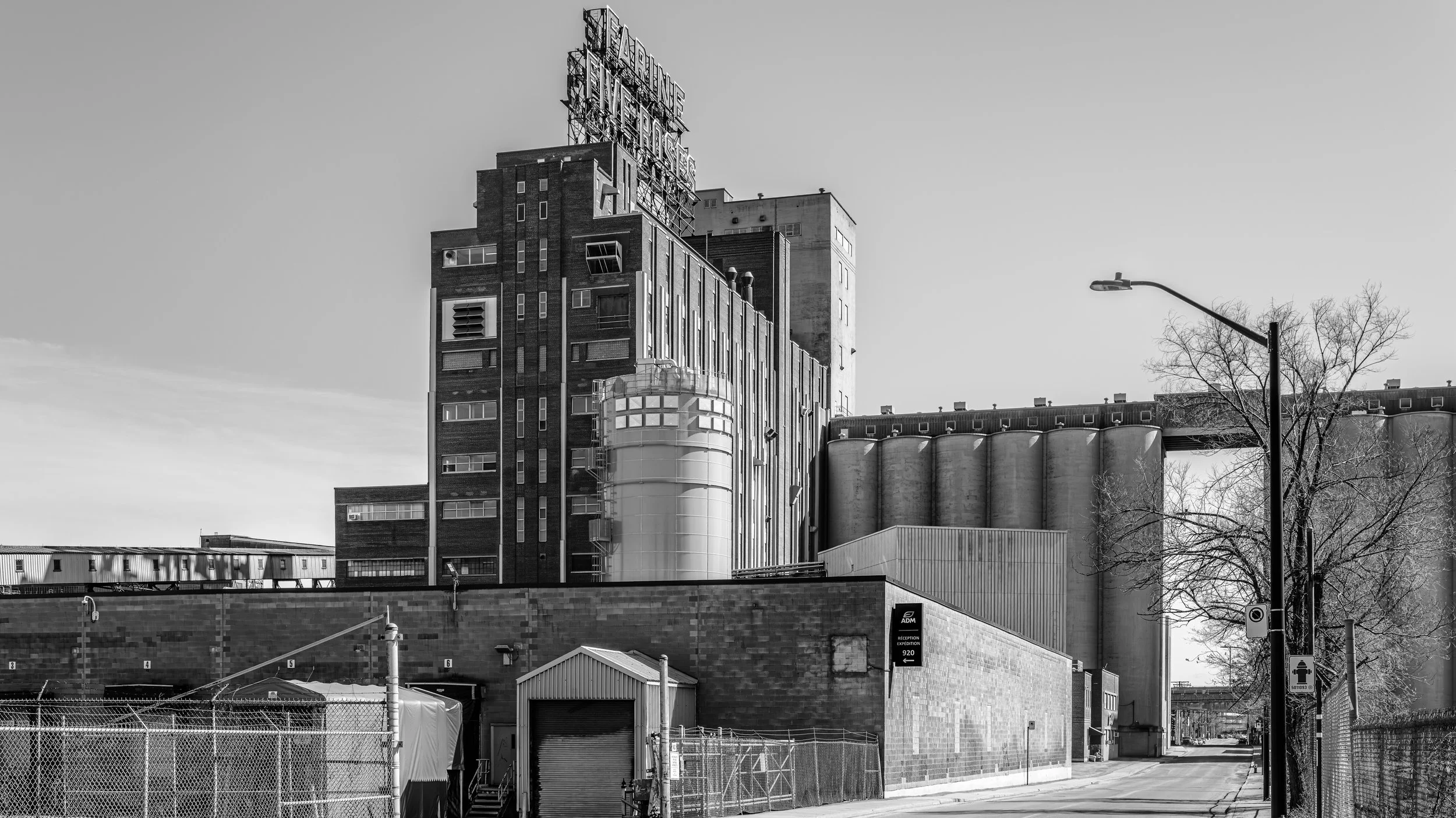 A black and white photo of an urban industrial building with a neon sign on top. The building has multiple windows and a rounded, glass-enclosed section. In the foreground, there are fences, a small structure, and a streetlamp, with a few leafless tr