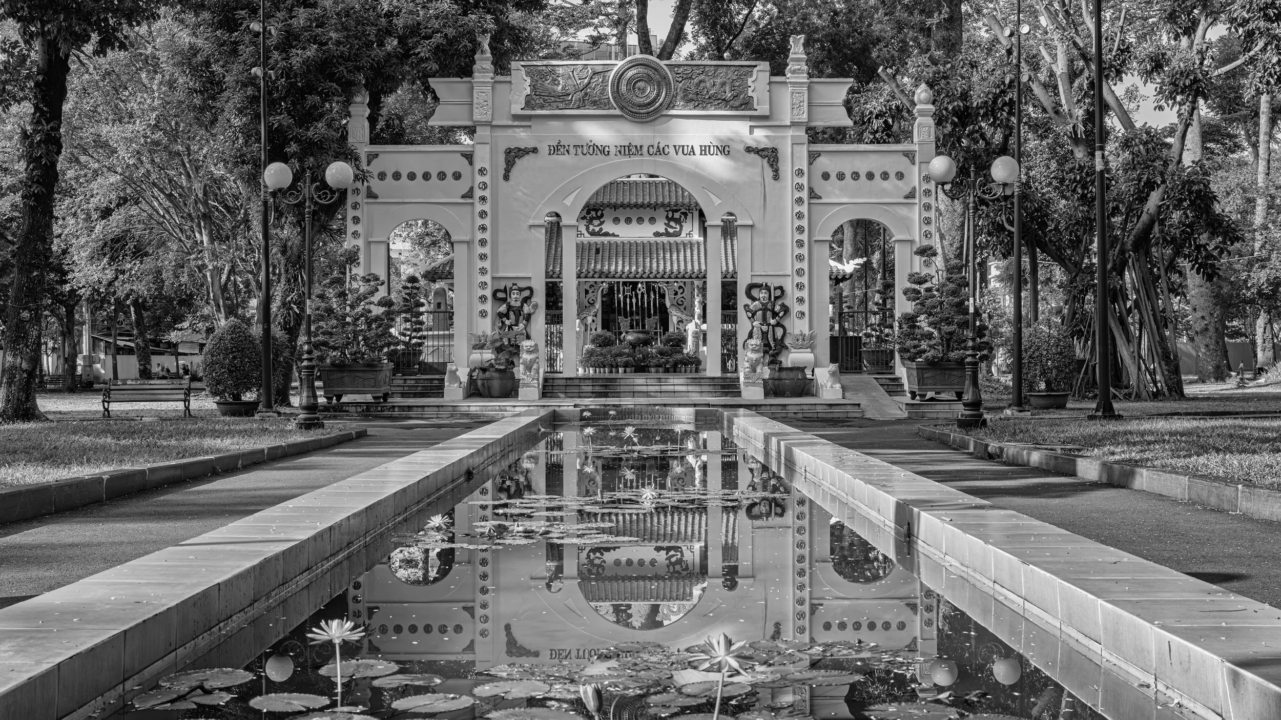 Grayscale photo of a decorative temple entrance with statues, a fountain, and reflections in water surrounded by trees and park benches.