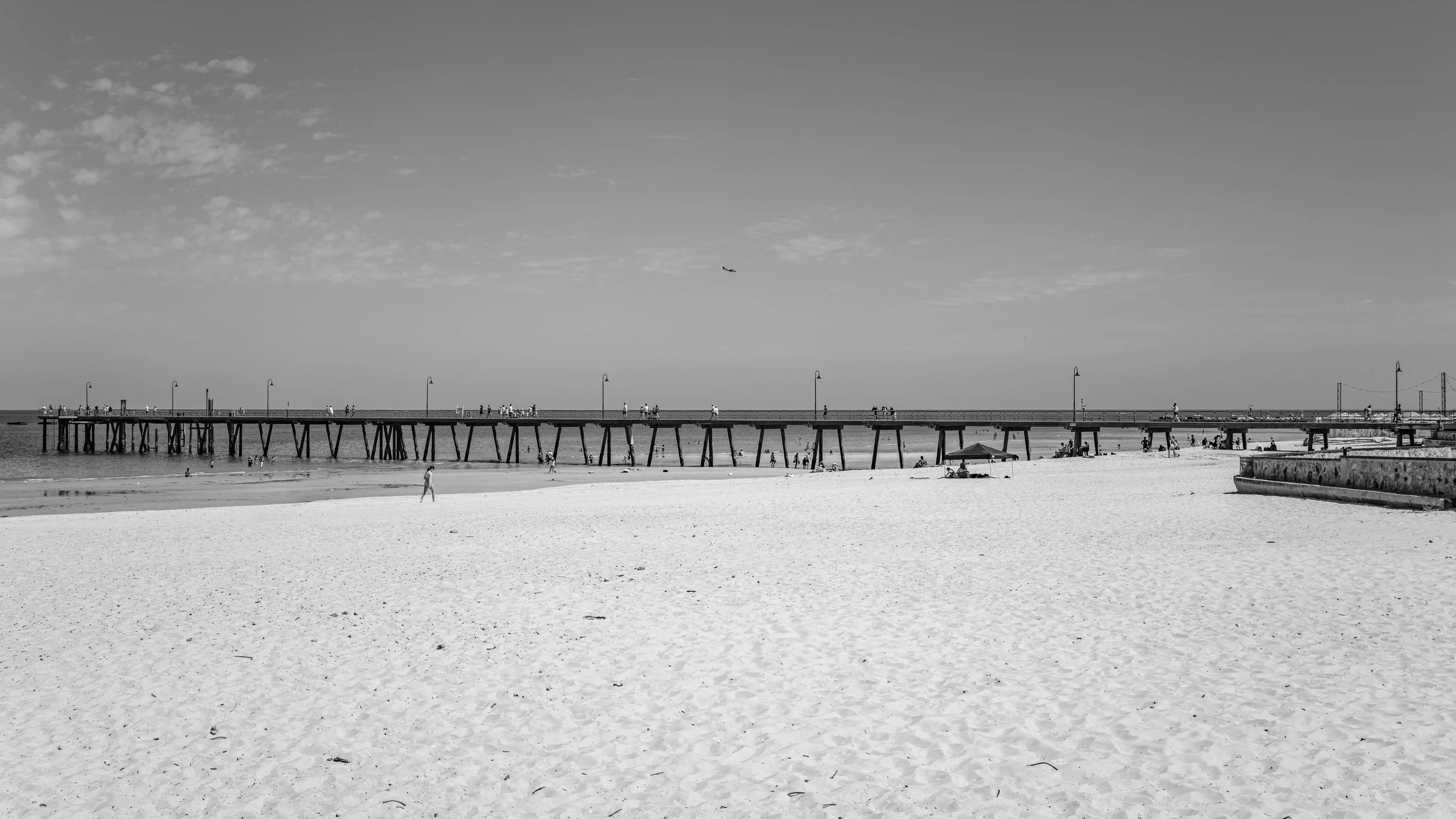A black and white photo of a beach with a pier extending into the water, people walking on the pier, some people sitting under an umbrella. Adelaide, Australia.