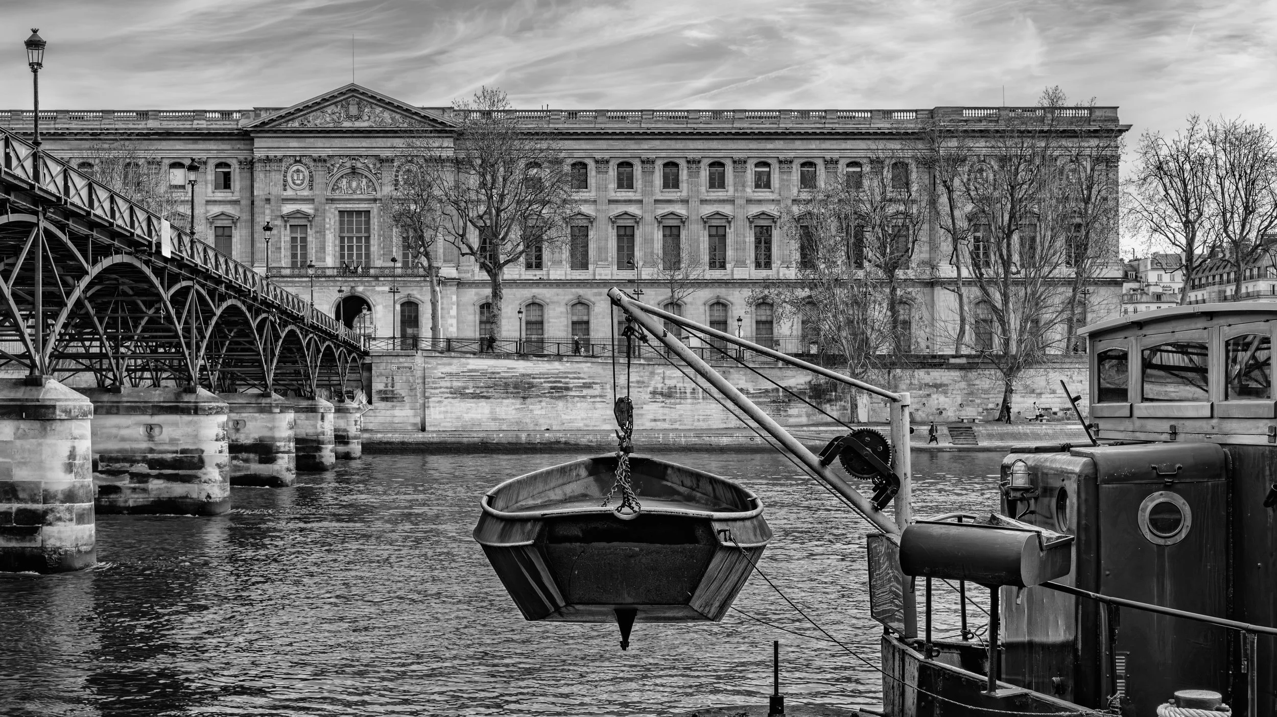 Black and white photo of a historic building along a river with a bridge on the left, trees, and boats in the foreground. Louvre Museum, Paris, France.