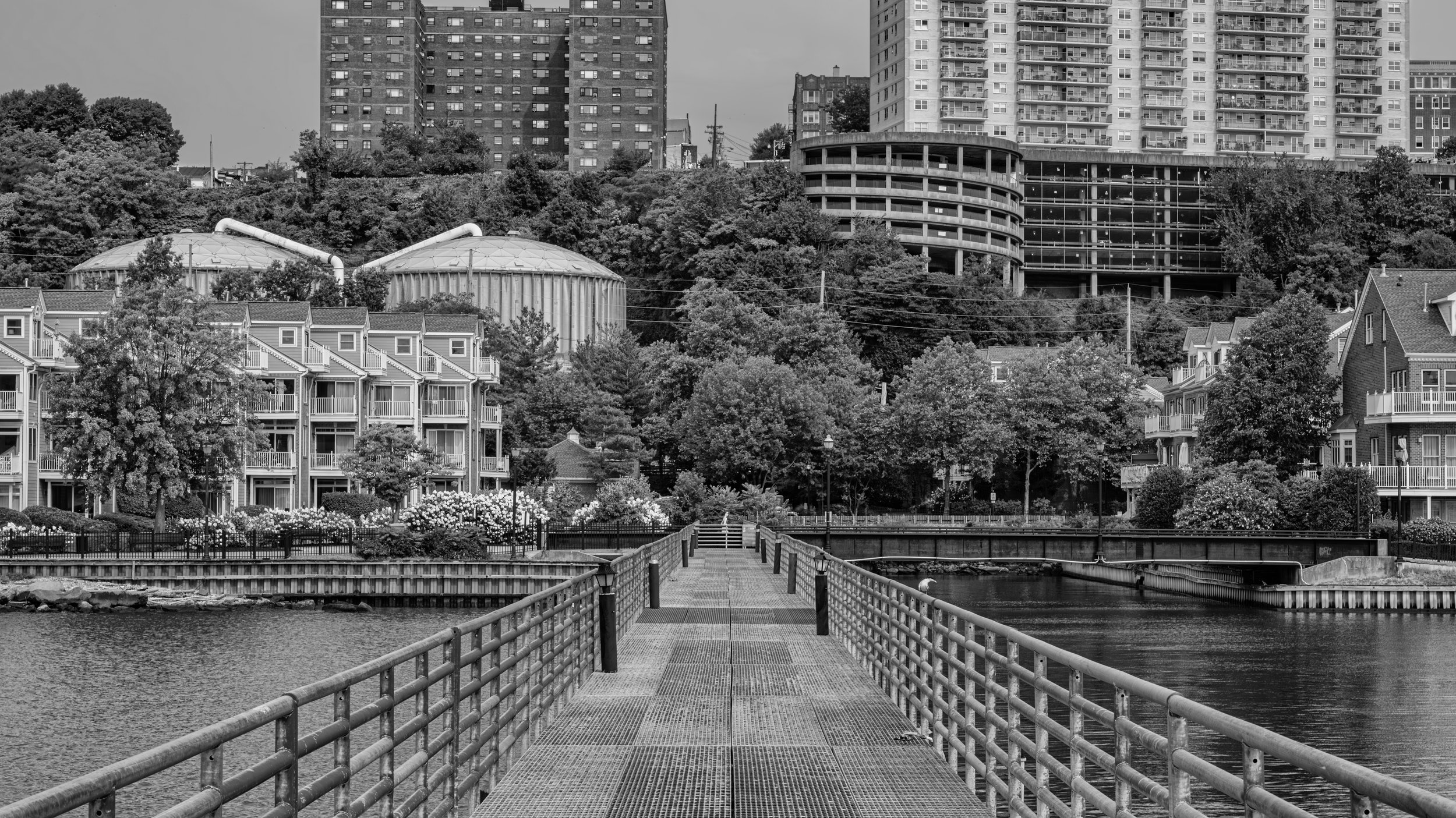 A black-and-white photo of a dock extending over water, leading to a cityscape with trees, residential houses with balconies, and large high-rise buildings in the background. Manhattan, New York, USA.