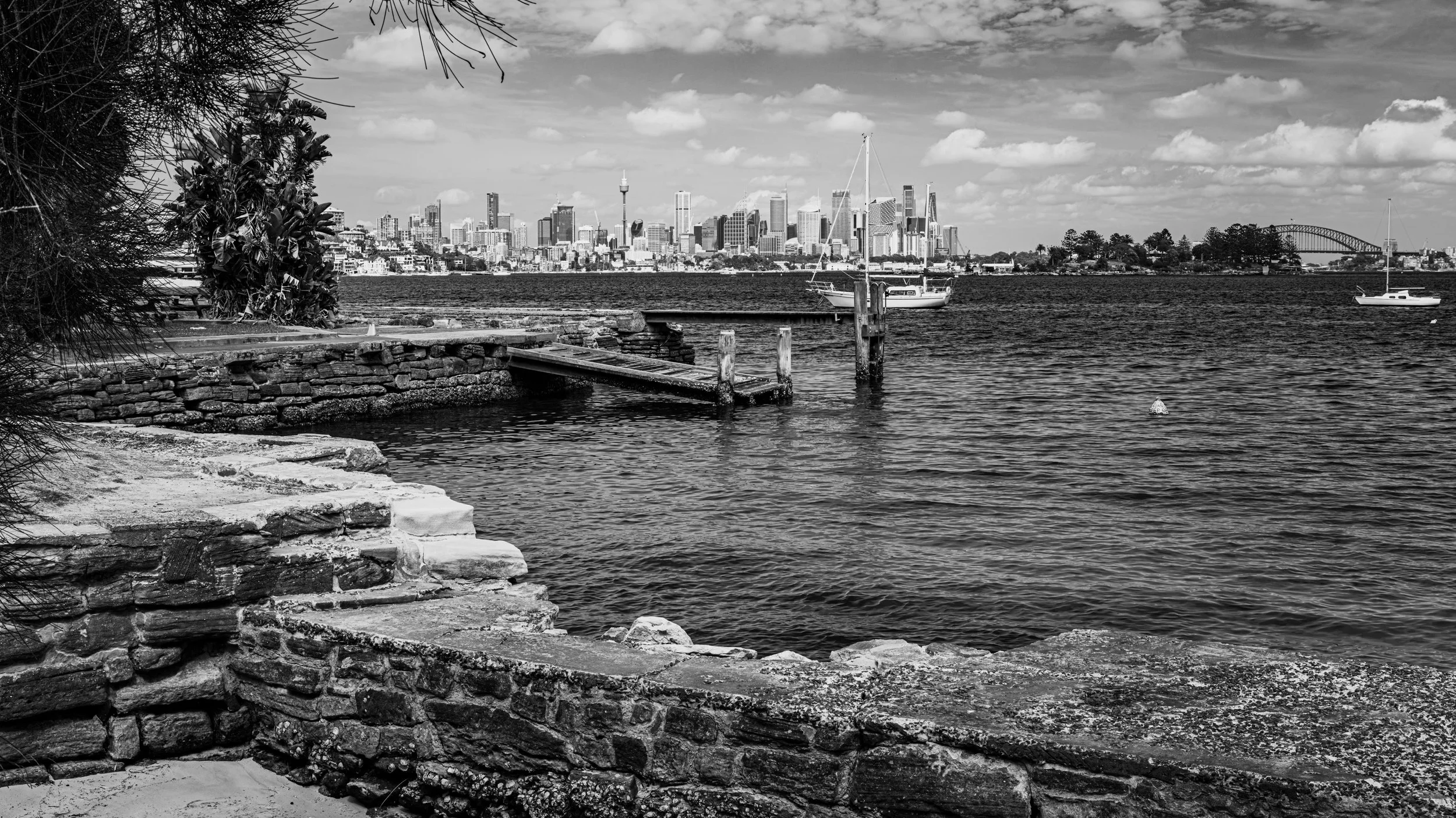 A black and white photograph of a waterfront with a city skyline in the background, including tall buildings, a tower, and a bridge; two sailboats are on the water, and there is a stone embankment and tree branches in the foreground.