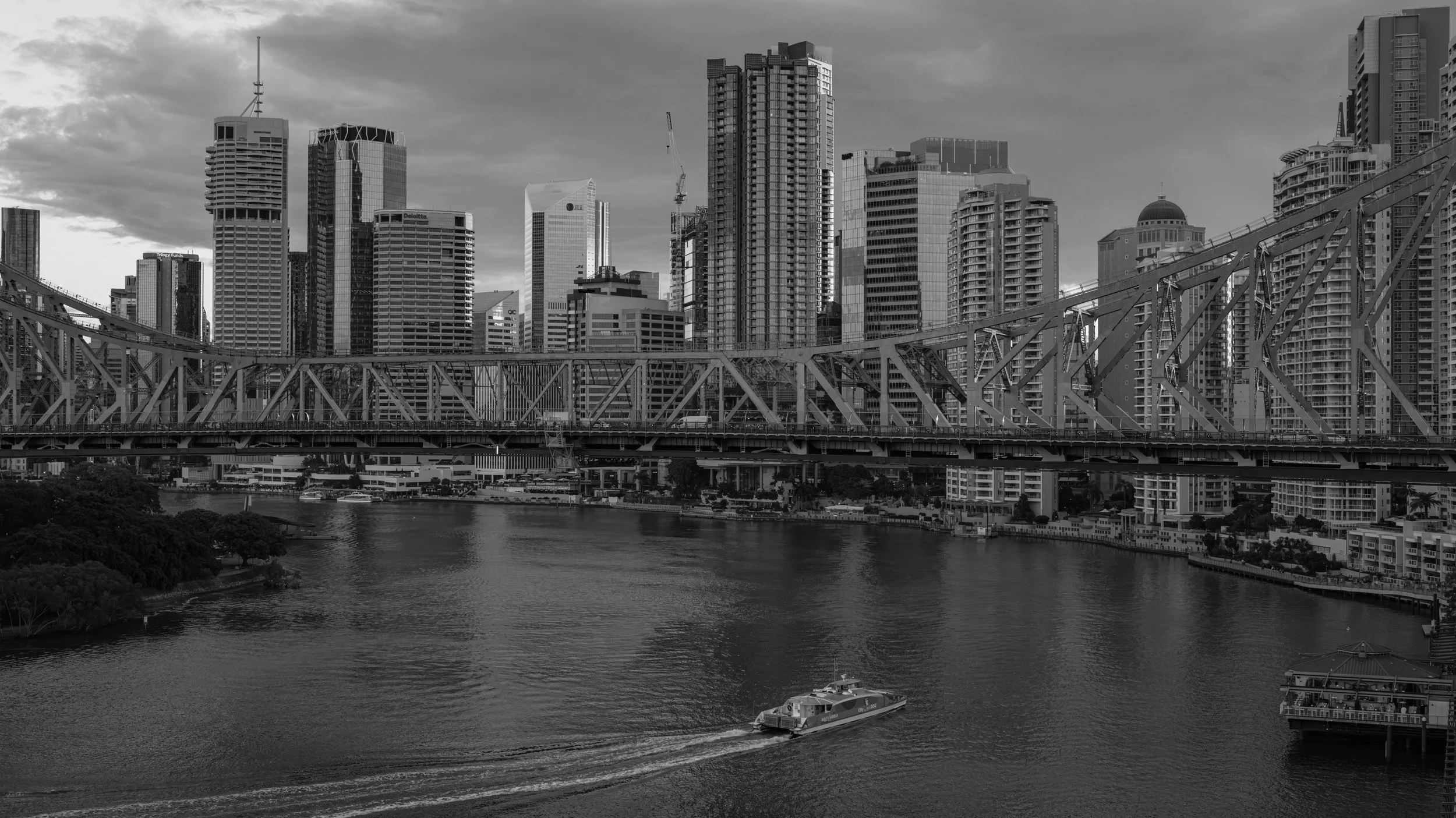 Black and white photo of a city skyline with numerous tall buildings and a bridge over a river, with a boat on the water.