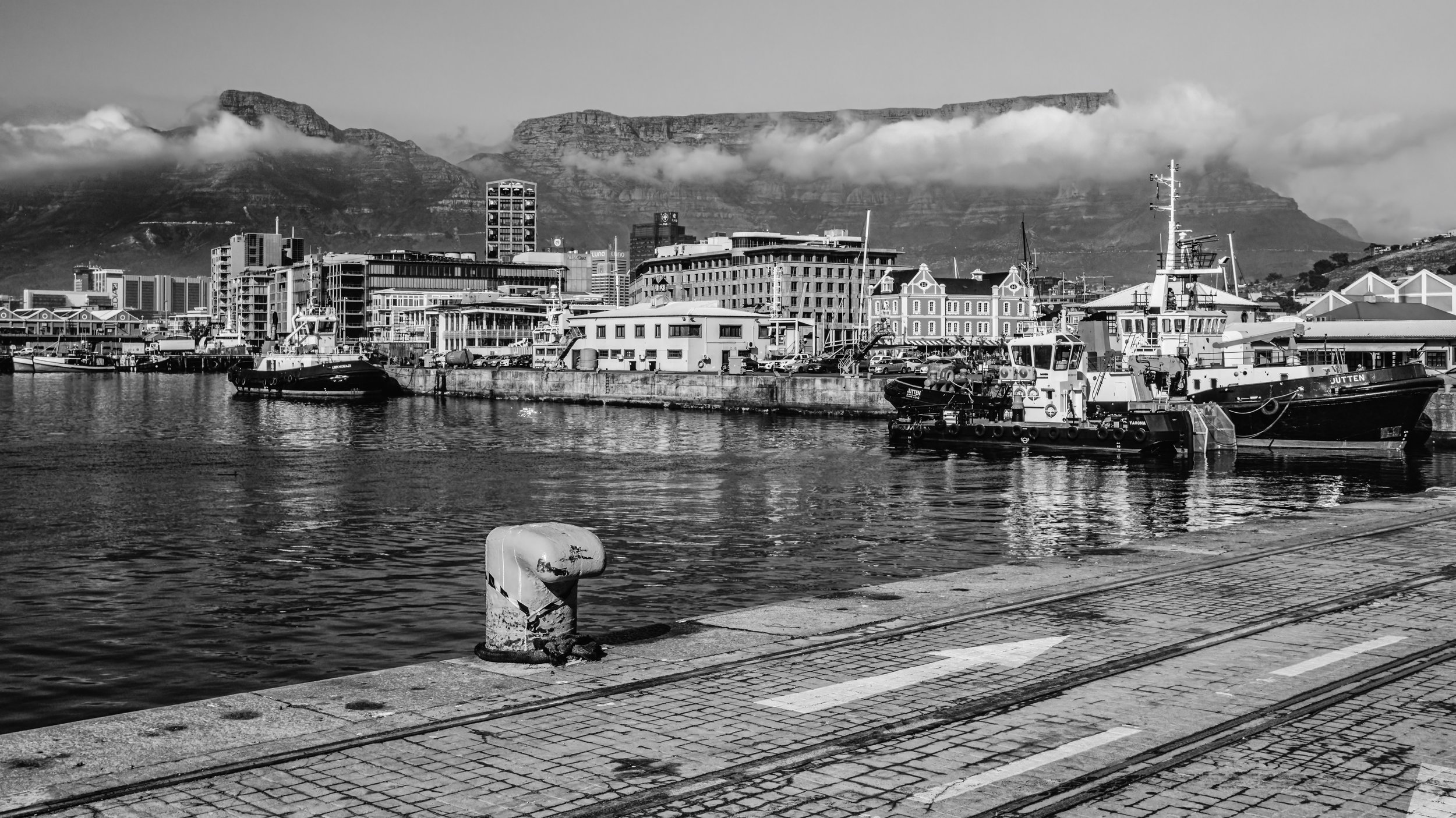 Black and white photograph of a harbor with boats docked, modern buildings in the background, and mountains with low clouds in the distance. Cape Town, South Africa.
