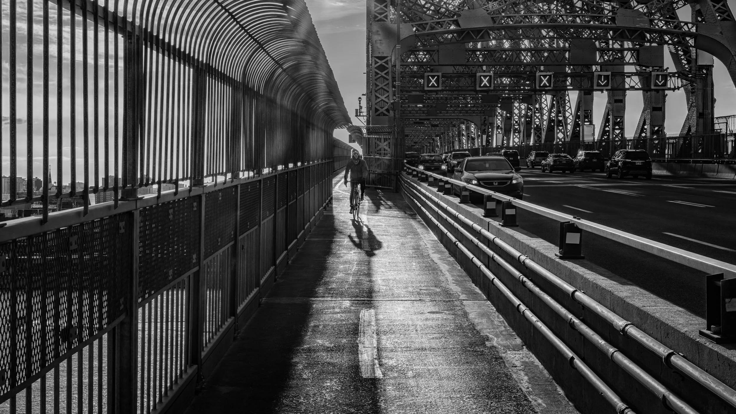 Black and white photo of a cyclist on a sidewalk next to a bridge with vehicles. The bridge has metal framework and structure, and a city skyline is visible in the background.