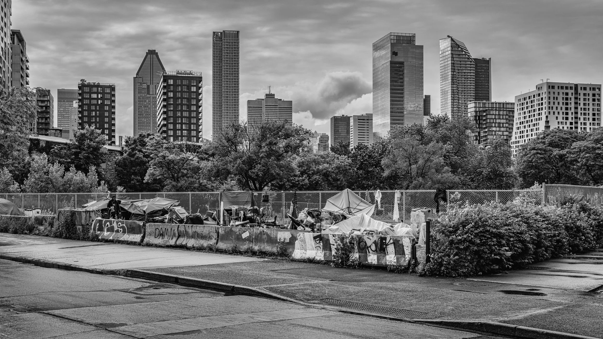 Black and white photo of a city skyline with tall modern buildings behind a fenced area filled with tents and debris, trees in the middle ground, and a sidewalk in the foreground.