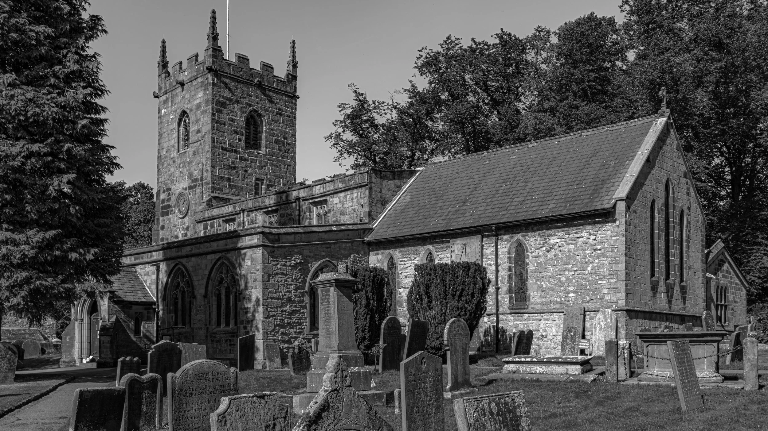 An old stone church with a tall square tower and arched windows, surrounded by a graveyard with headstones, trees, and a cloudy sky in black and white.