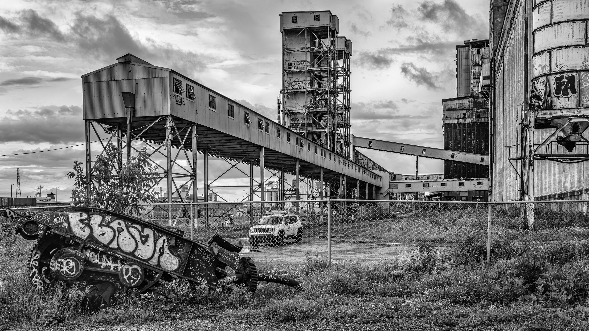 Black and white photo of an industrial area with old factories, silos, and a metal bridge, surrounded by a chain link fence, with graffiti on a toppled vehicle in the foreground and a cloudy sky overhead. Montreal, Canada.