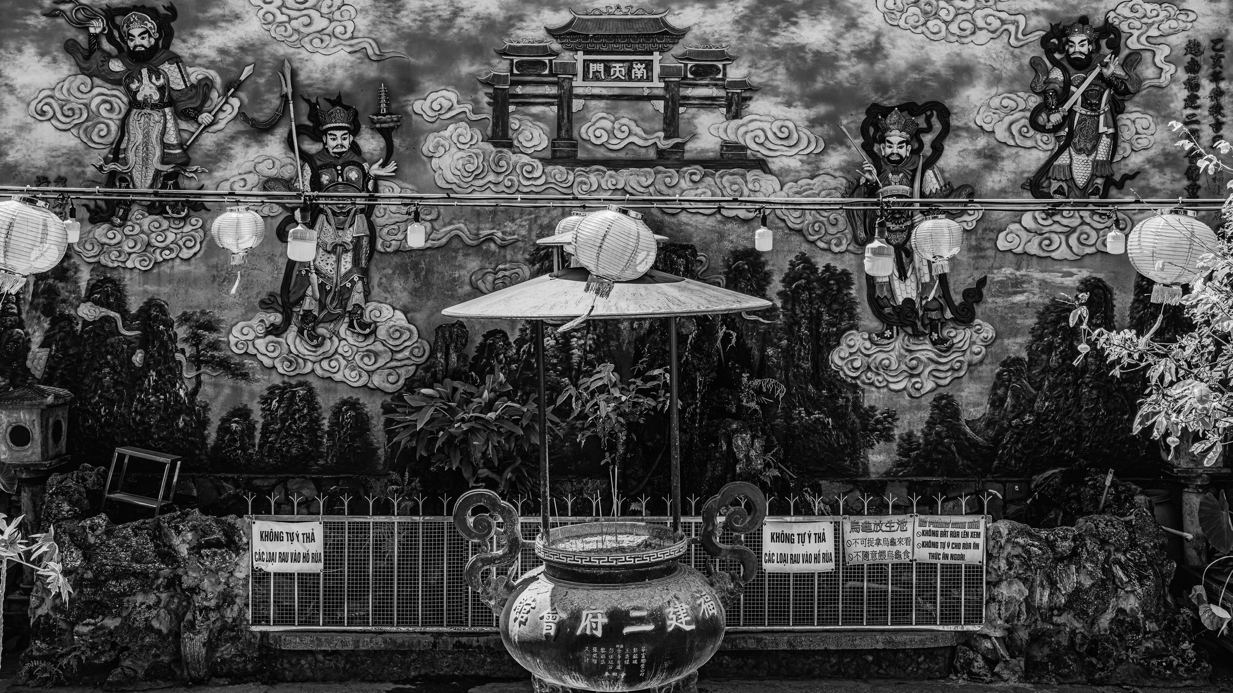 A black-and-white mural featuring four mythological Chinese warriors on clouds above a landscape with trees. In front, a large ornate urn and a sign with Chinese and Vietnamese text are visible, along with paper lanterns hanging across the scene.