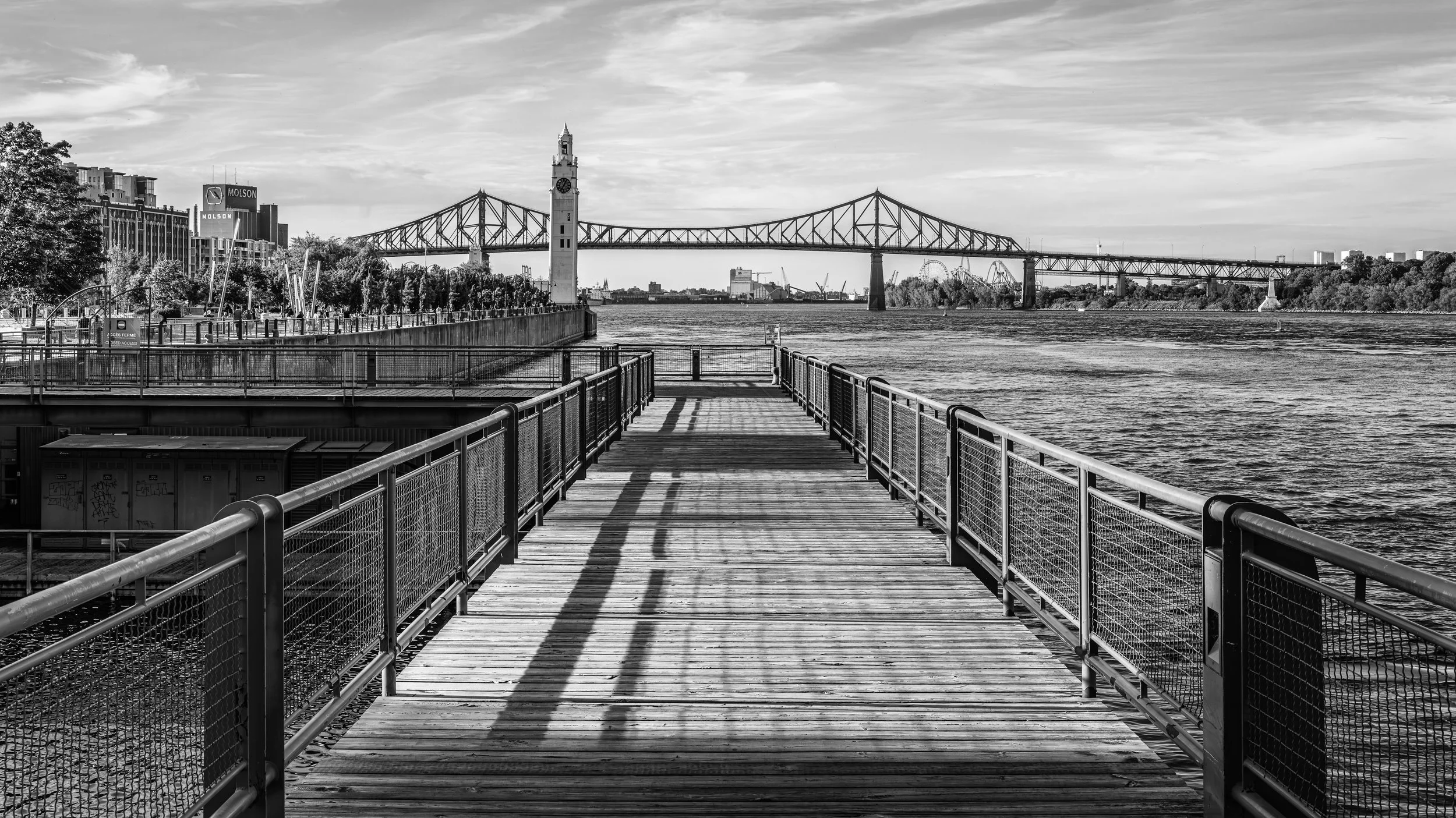A black and white photo of a waterfront scene featuring a pier with railings, a clock tower, a large bridge in the background, and a city skyline with buildings and a Ferris wheel.