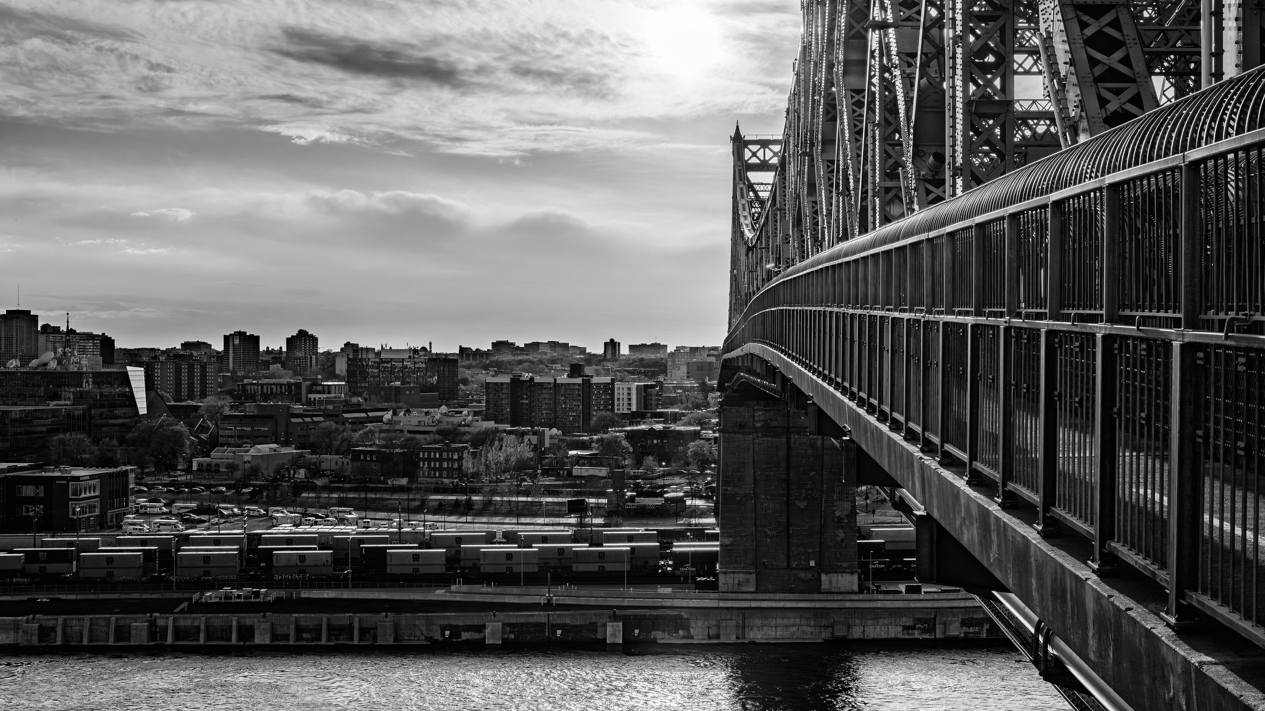 Black and white photo of a city skyline with a large bridge on the right side, spanning over a body of water, with numerous buildings in the background under a cloudy sky.