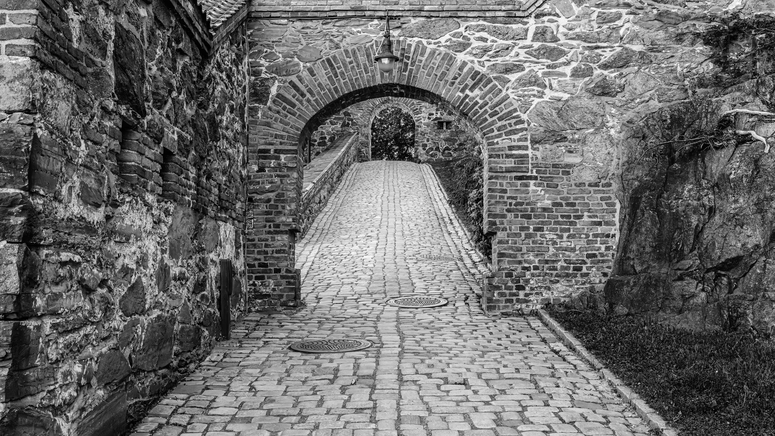 Black and white photo of a narrow cobblestone alleyway with brick and stone walls on either side, leading to an arched brick passageway with a view of trees in the distance.