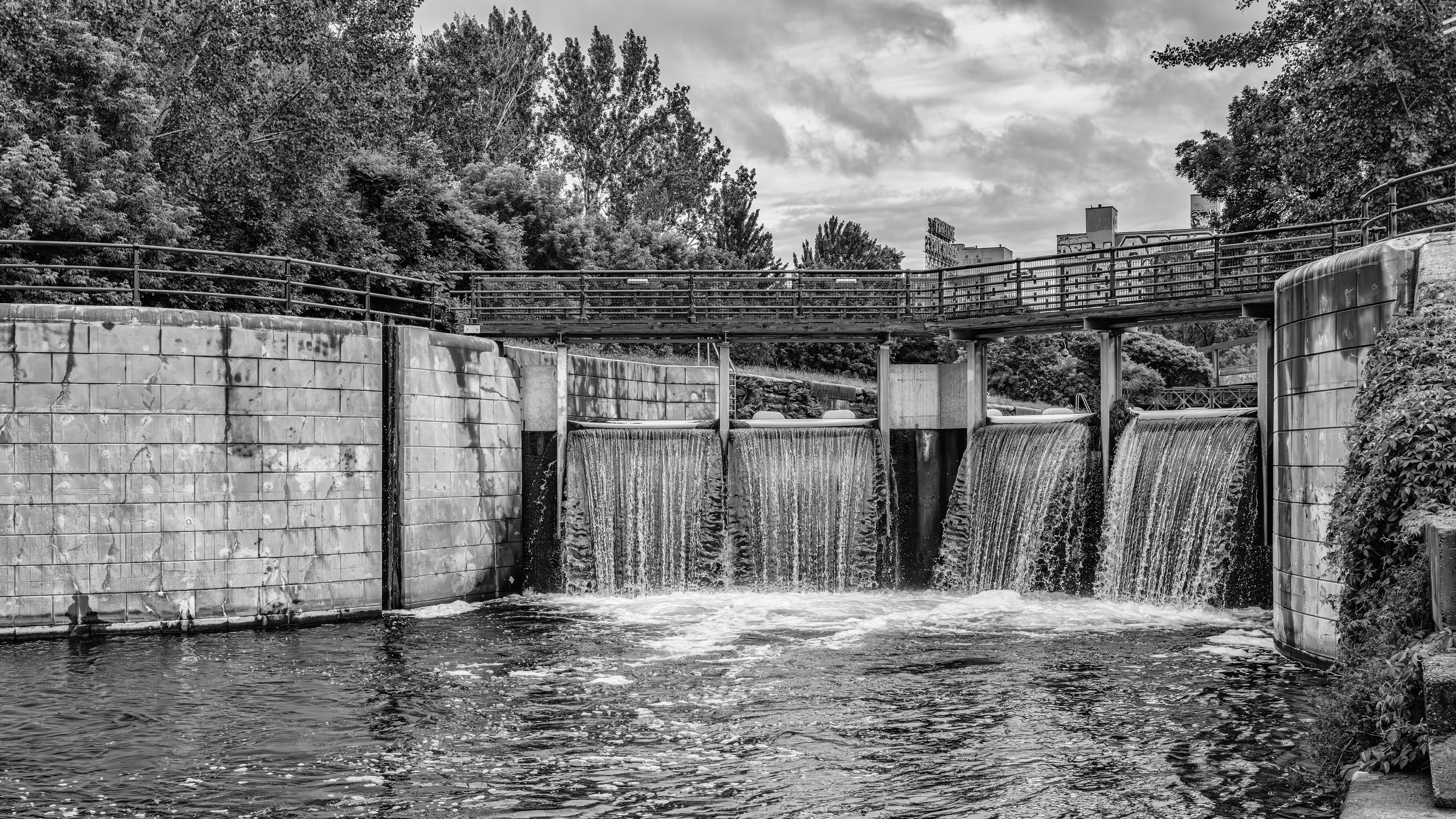 A black-and-white photo of a dam with water flowing over the edge, trees in the background, and a cloudy sky.