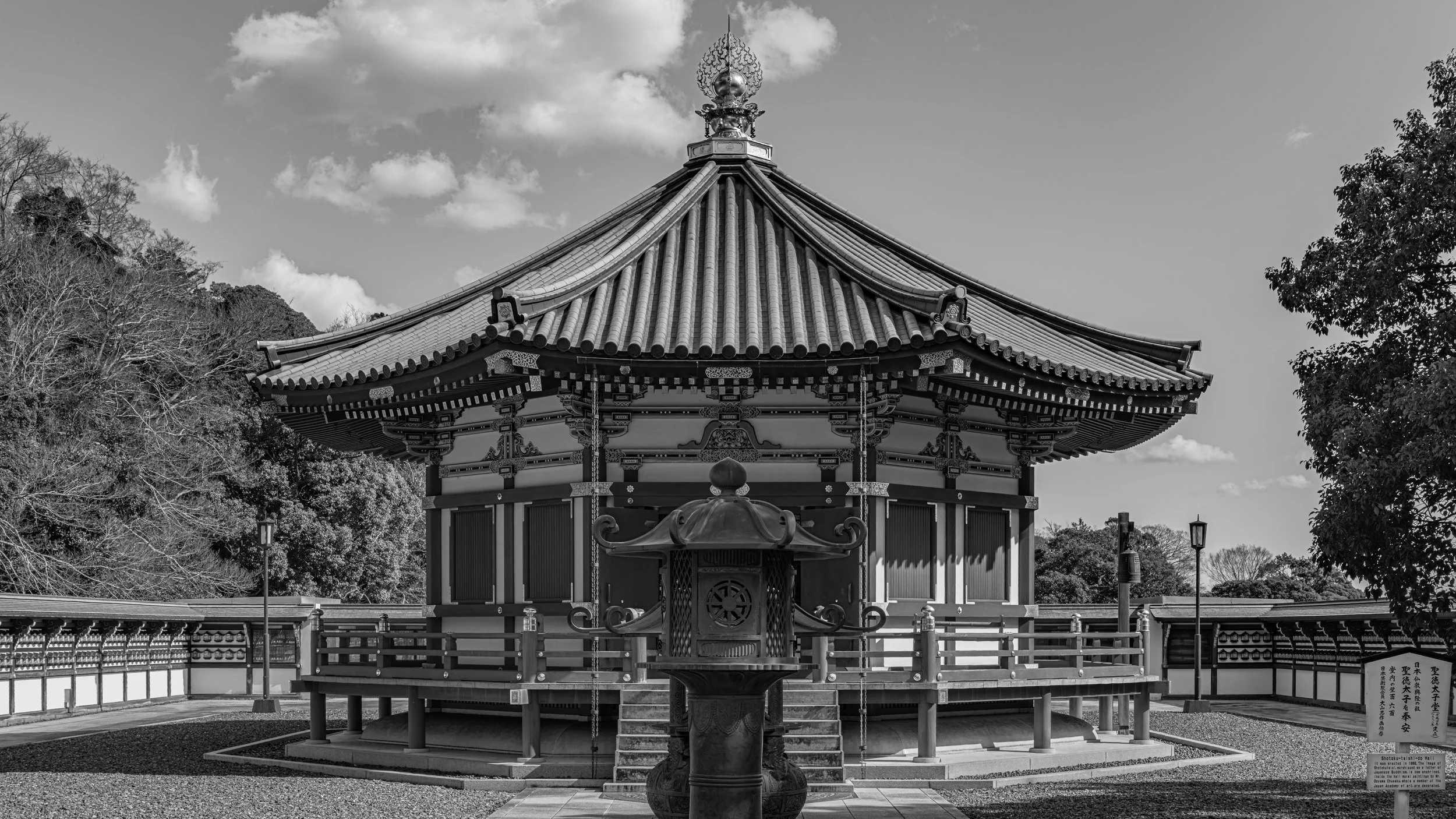A traditional Japanese pagoda with ornate roof tiles, surrounded by trees, under a partly cloudy sky, in black and white.