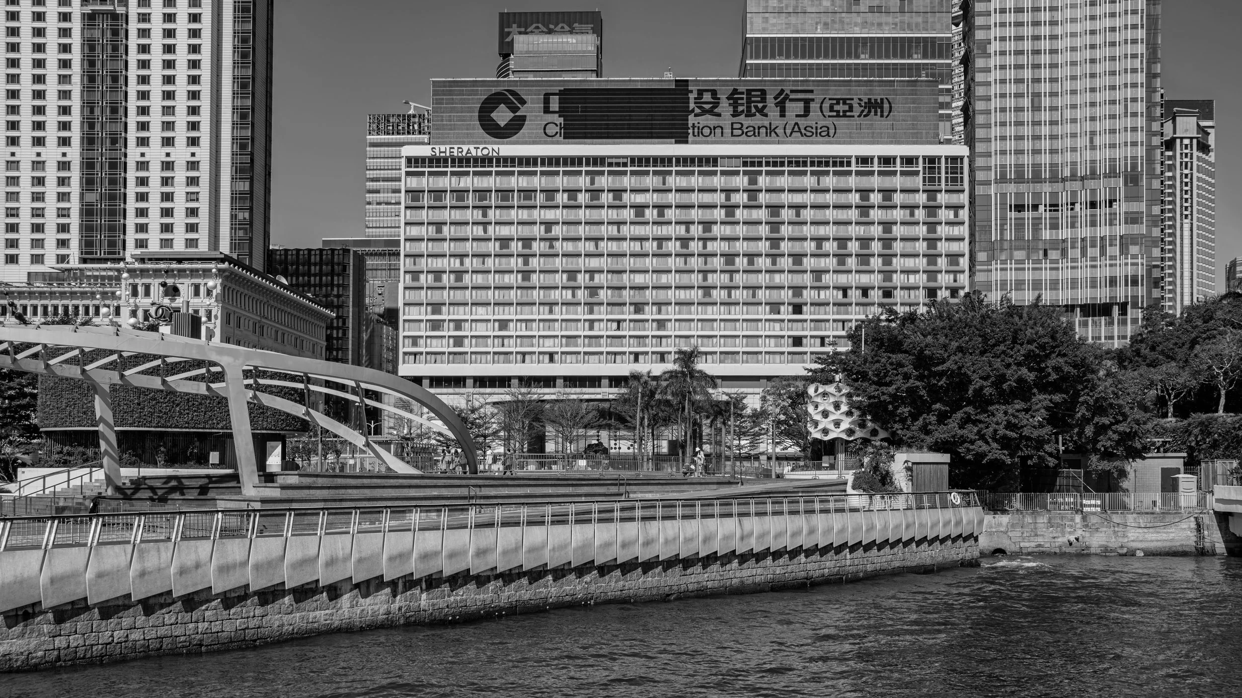 Black and white photo of a city skyline with tall modern buildings, a waterway in the foreground, and trees near the riverbank.