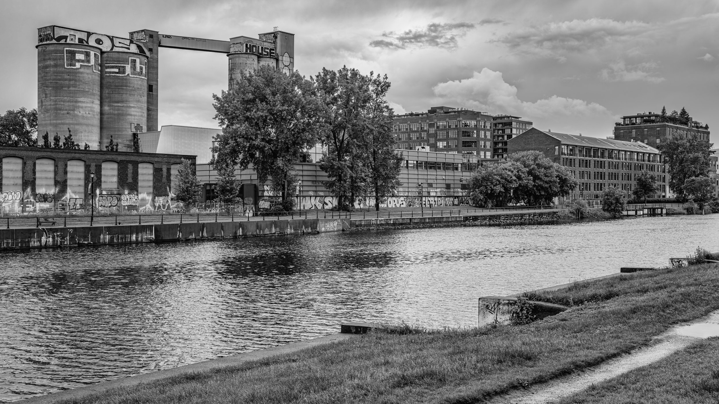 Black-and-white photo of an urban waterfront with buildings, trees, and industrial silos, with cloudy sky above.