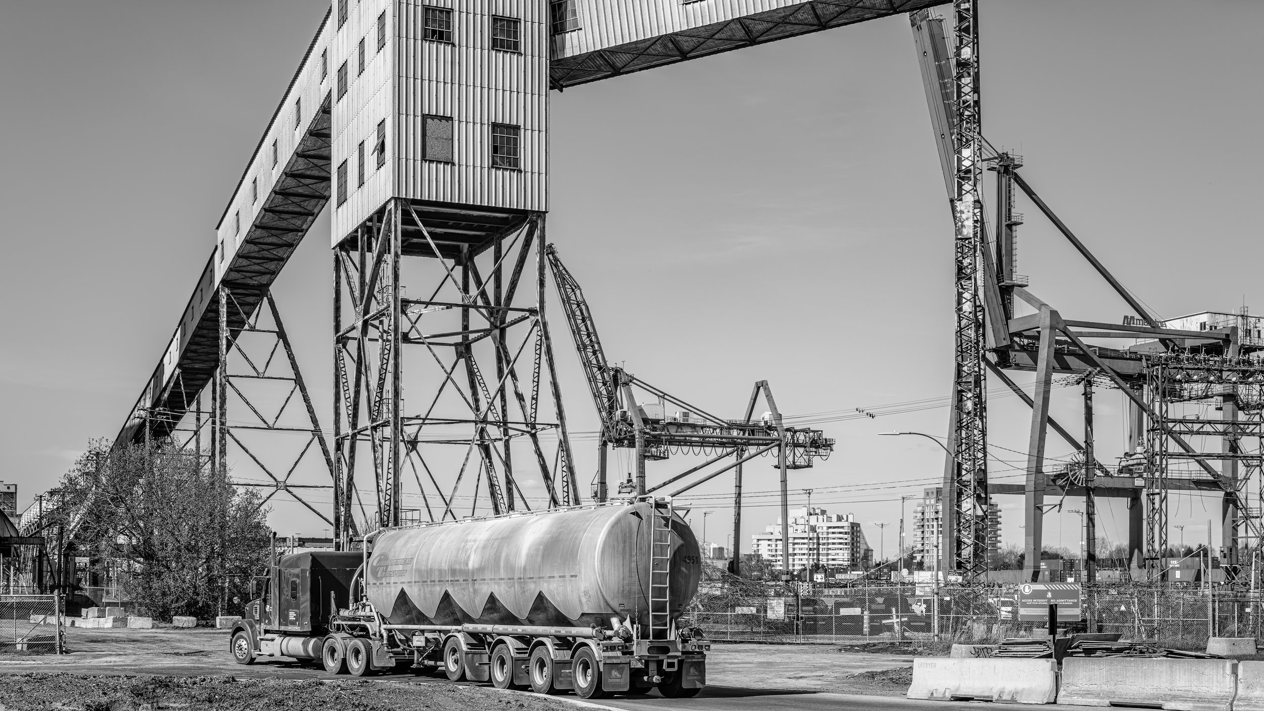 Black and white photo of an industrial construction site featuring large steel structures, cranes, and a tanker truck.