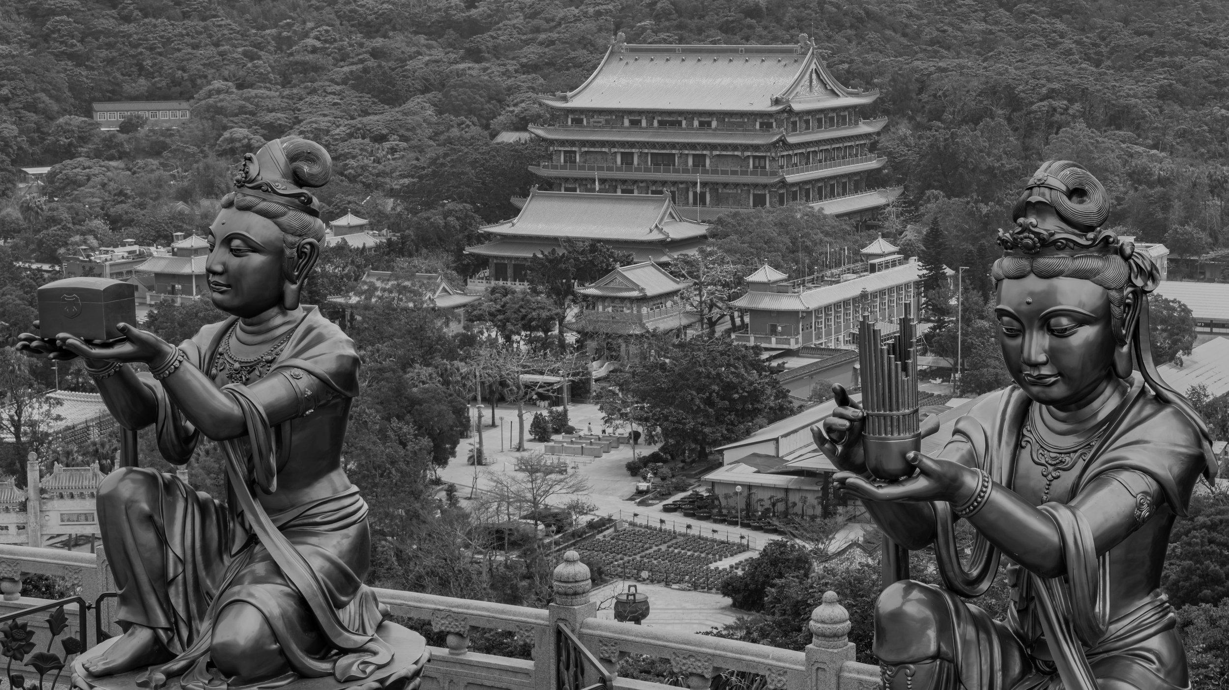 Black and white photo of two ornate statues of women holding objects in front of a traditional East Asian temple complex with multiple tiers, surrounded by trees.
