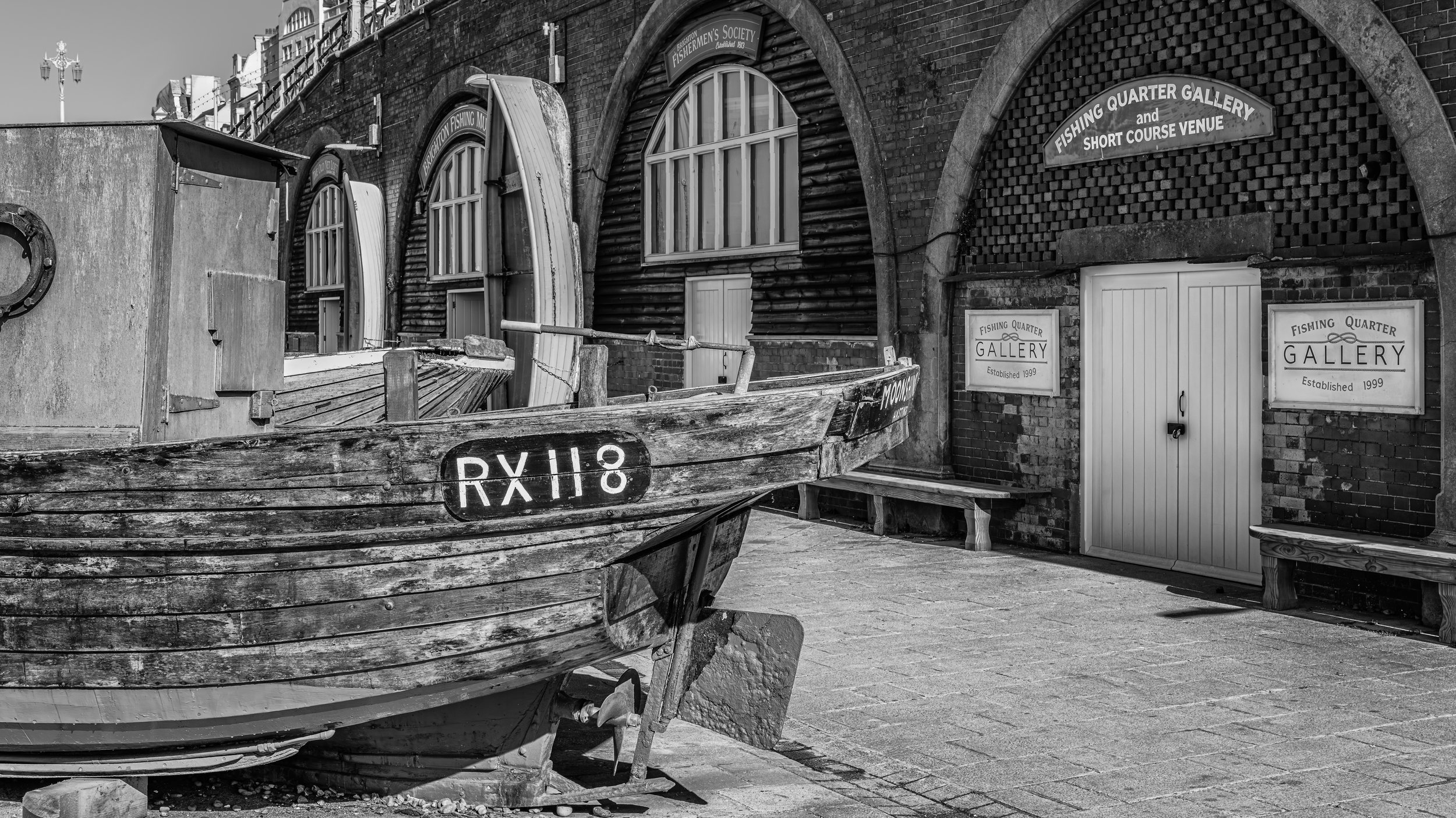 A black and white photo of a fishing boat with the registration number RX118 lodged against a brick building. The building has signs indicating it is a gallery and a short course venue, associated with a fishing quarter gallery established in 1999. T