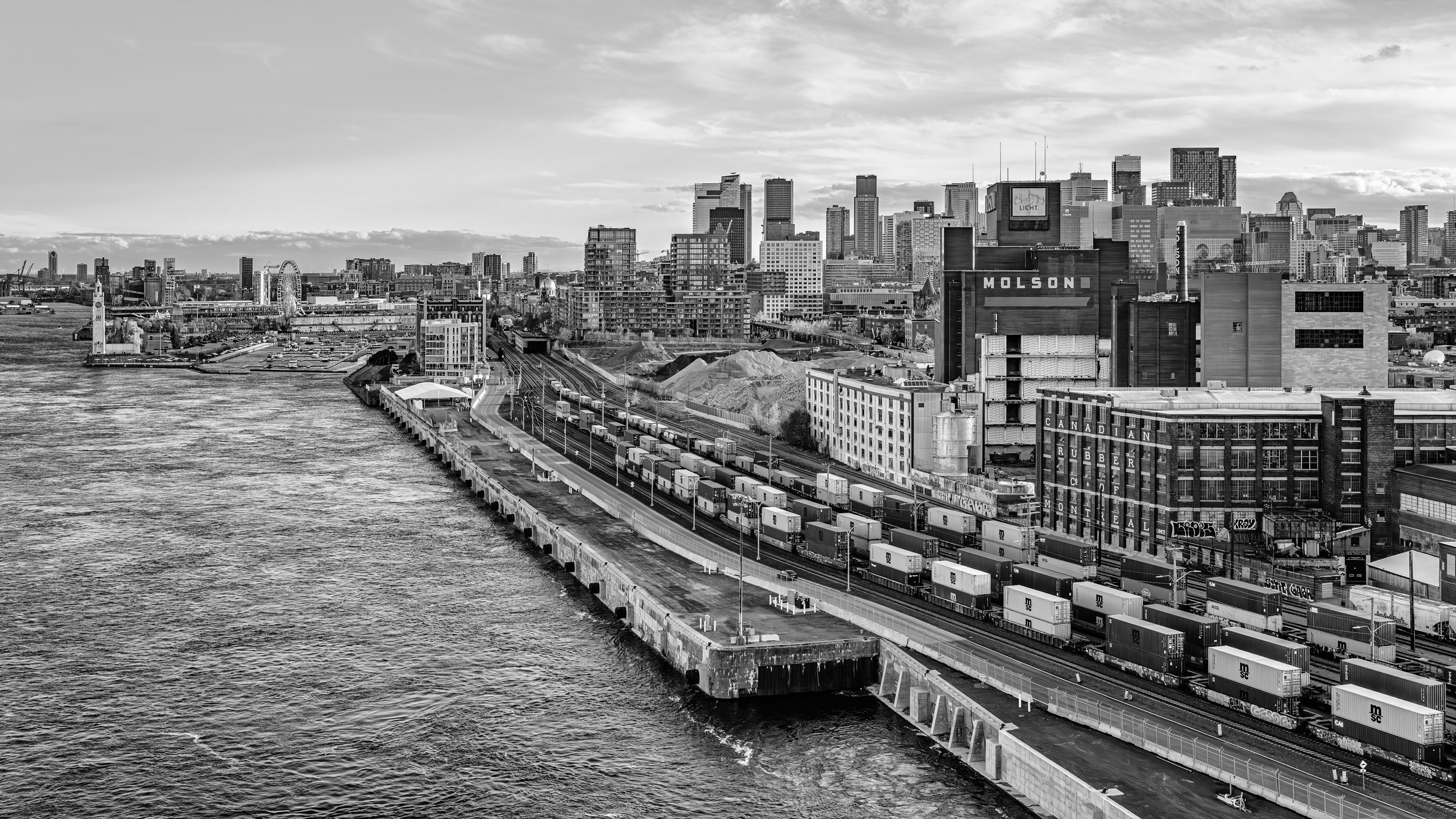 Black and white aerial view of a city skyline with tall skyscrapers, a waterway on the left, and a railway lined with freight containers in the foreground.