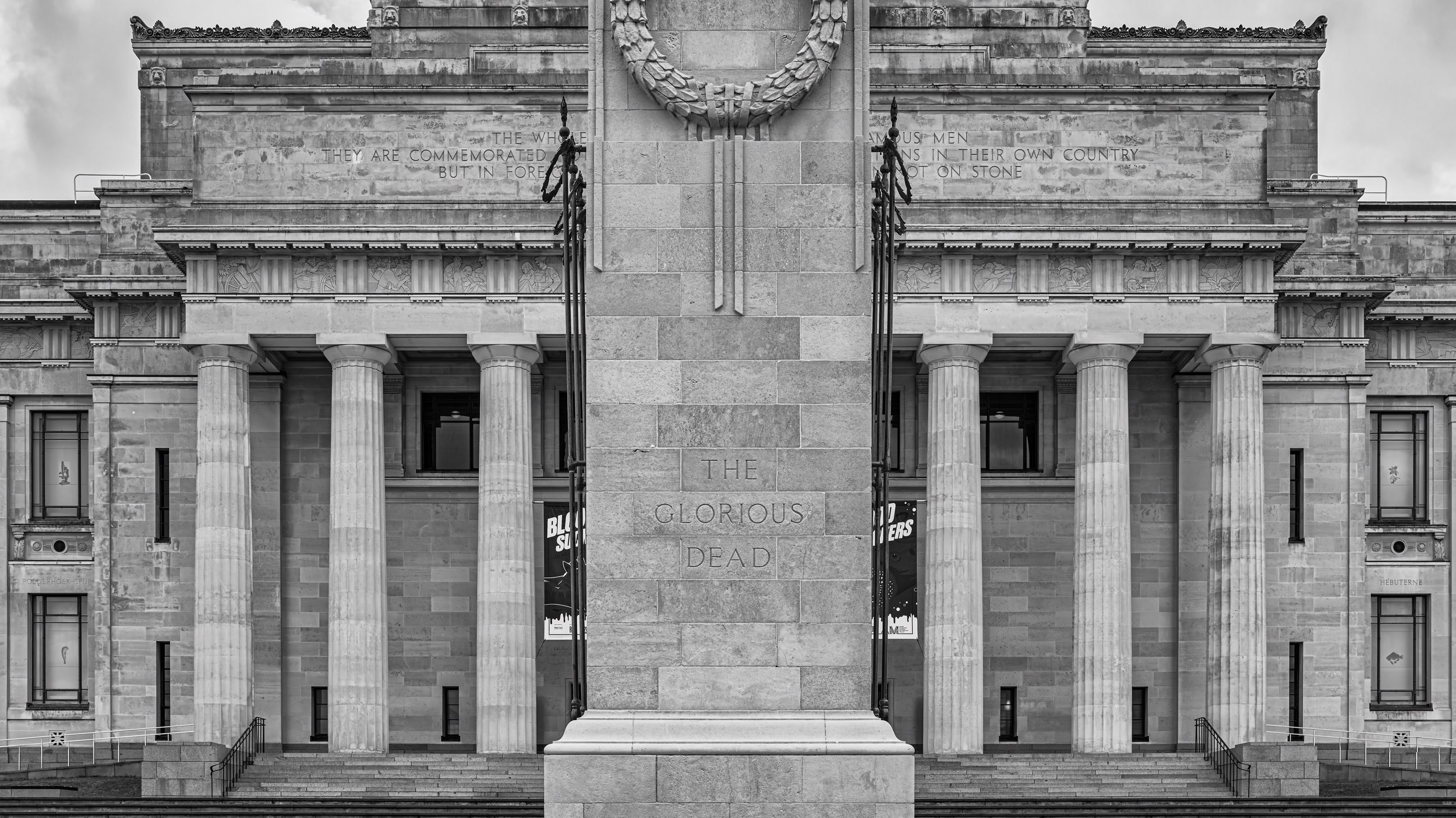 Black and white photo of a large classical-style building with tall columns and engravings, featuring a central stone pillar with the inscription "THE GLORIOUS DEAD" and a large wreath sculpture above it.