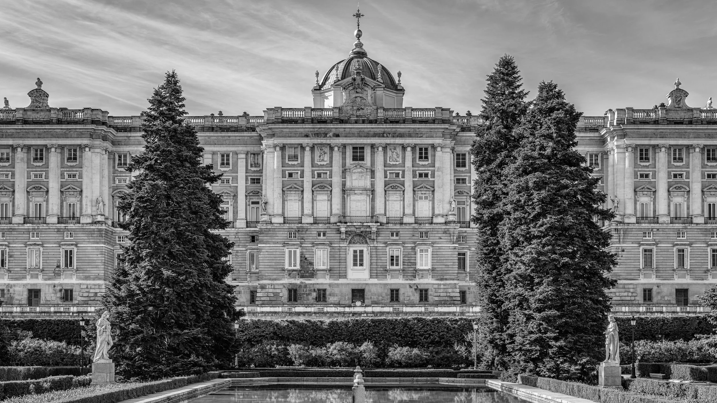Black-and-white photograph of a grand baroque-style palace or government building with a prominent dome and decorative statues on the roof, surrounded by tall, full trees and statues in the foreground.