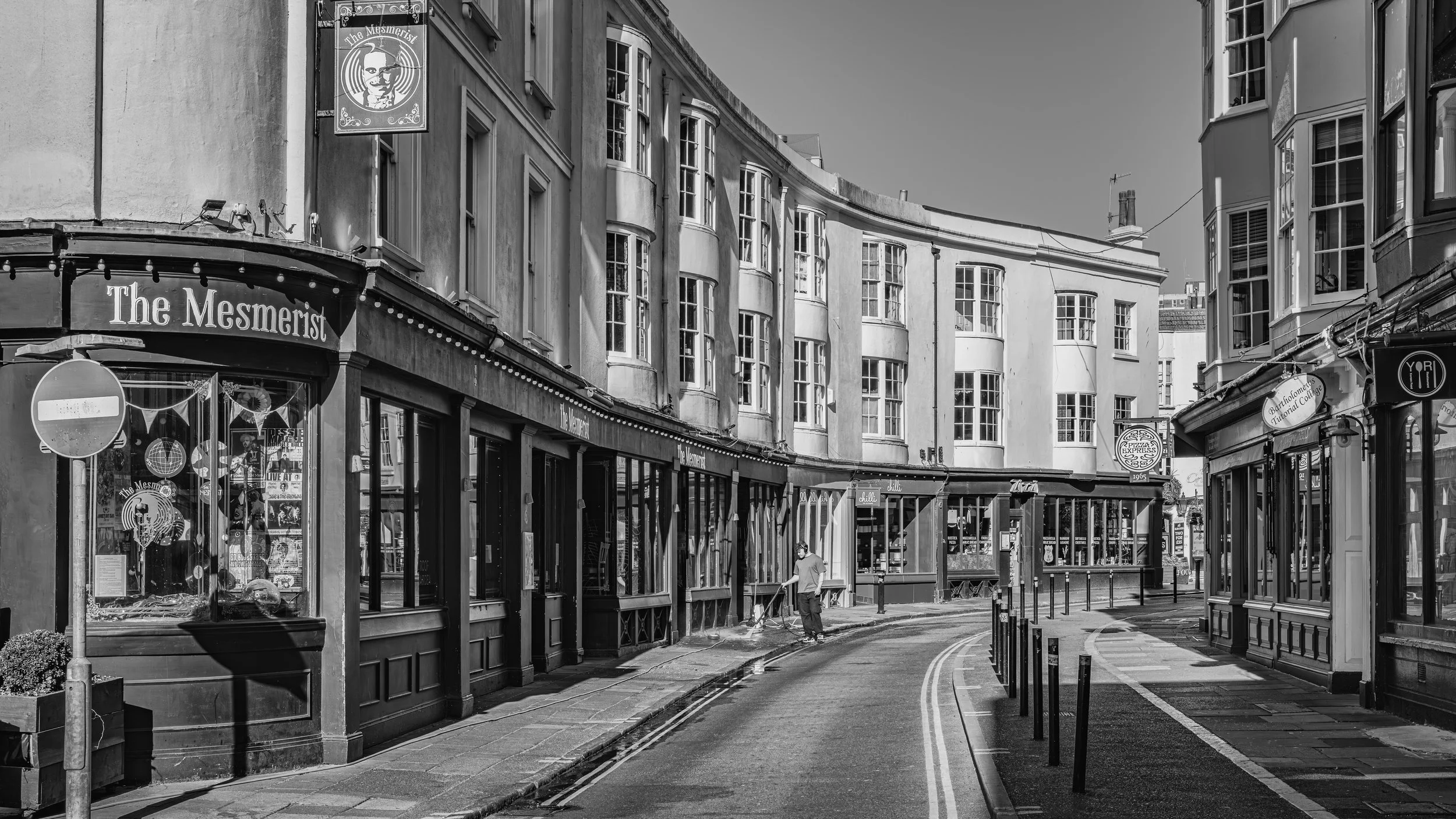 A black and white photo of a street scene with residential and commercial buildings. There is a sign for "The Mesmerist" on a building on the left and a street cleaner working in the middle of the street. The street curves to the right and is empty of cars and pedestrians.