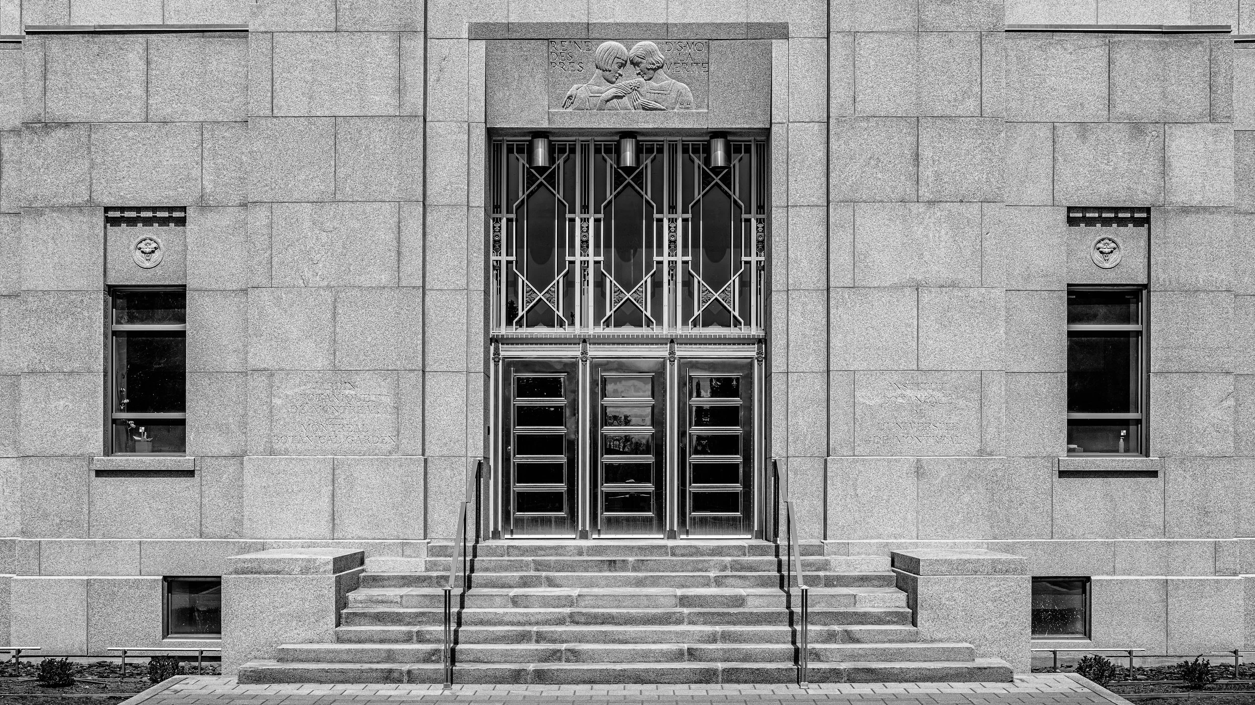 Black and white photo of the front entrance of a building with steps leading up to it. The building has a large metal door, two small windows on either side, and a decorative stone relief of two children above the door. Stone walls surround the entra