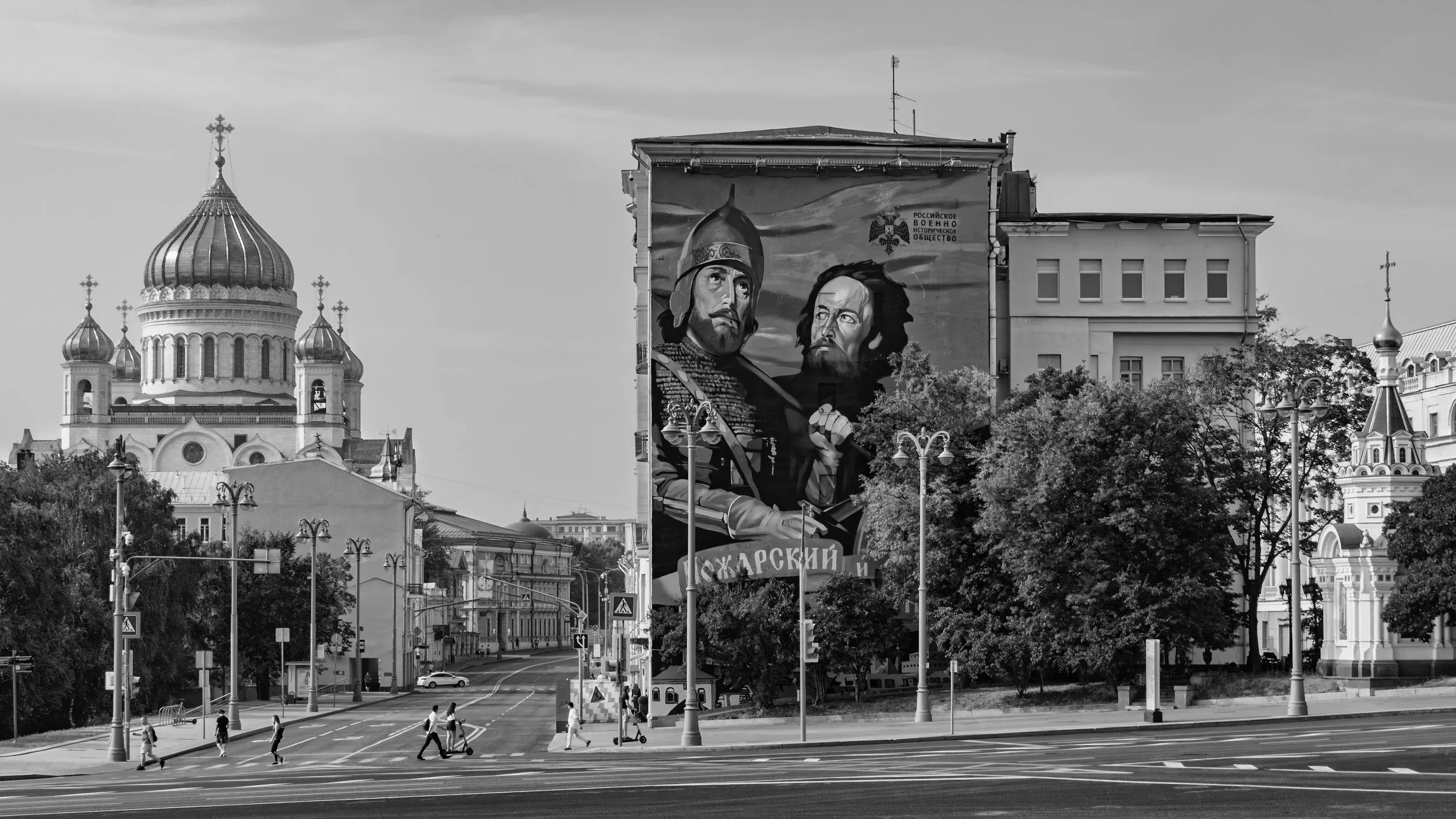 A cityscape with a large church with onion domes on the left, a mural on a tall building depicting two historical figures wearing helmets, trees, street lamps, and pedestrians crossing the street in the foreground. Moscow, Russia.