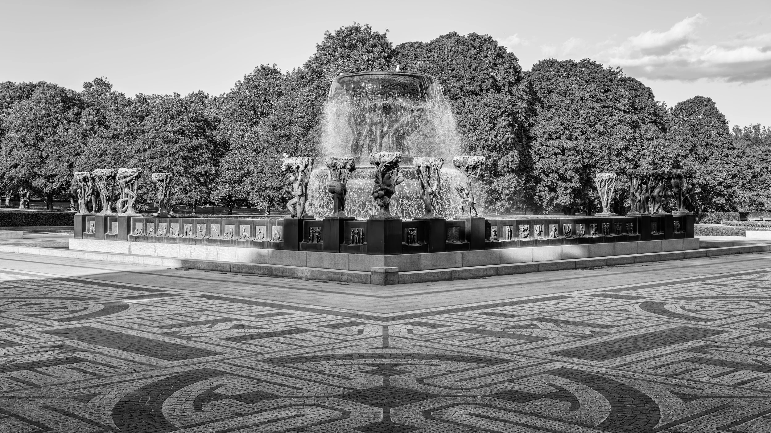 A large decorative fountain with sculptures of human figures and animals, surrounded by a paved area with intricate patterns, in a park with many trees in the background, under a partly cloudy sky.