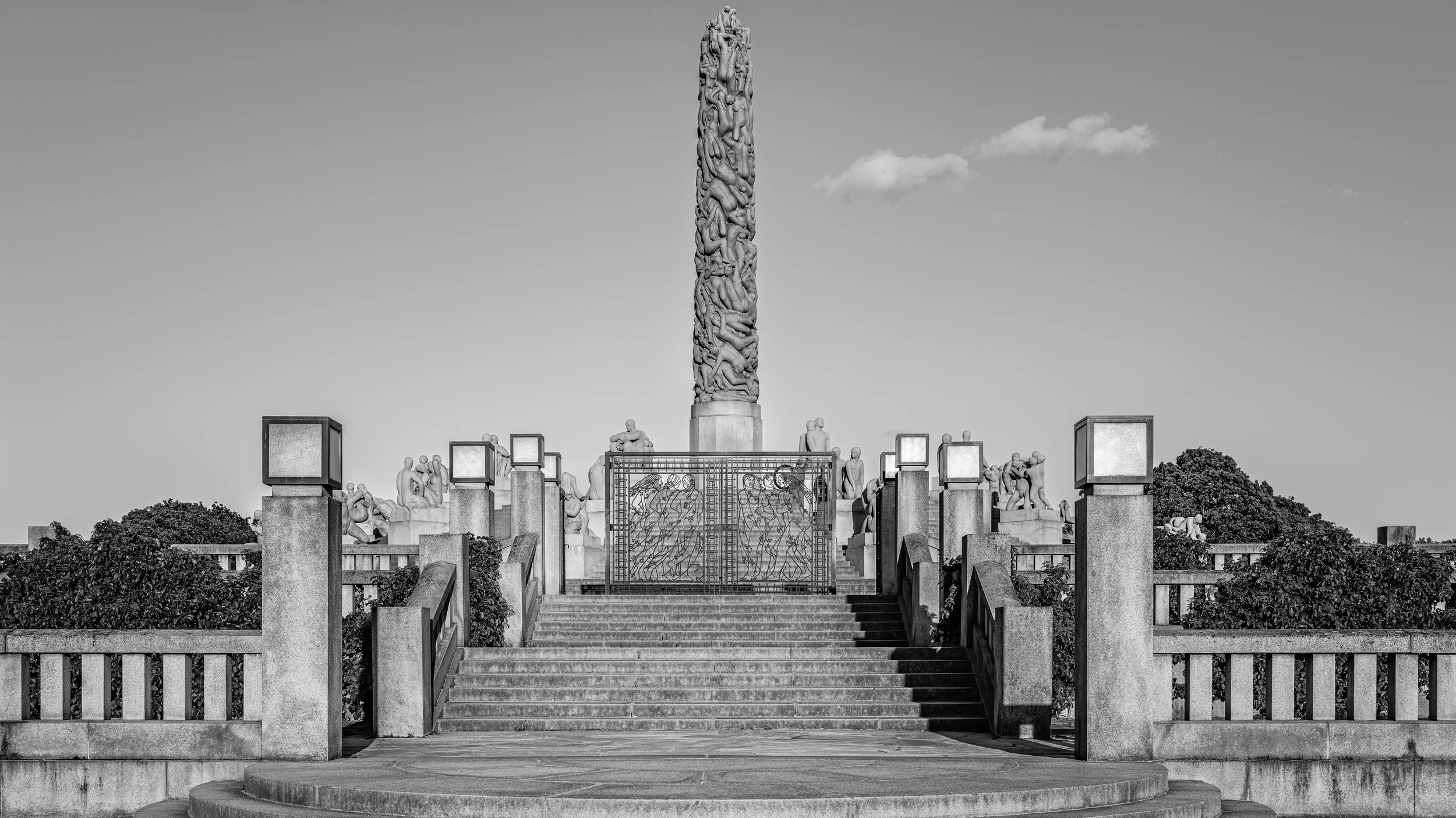 A monument with a tall, detailed sculpture column at the center, surrounded by numerous smaller sculptures of human figures, and stairs leading up to the sculptures across a landscaped area with trees.