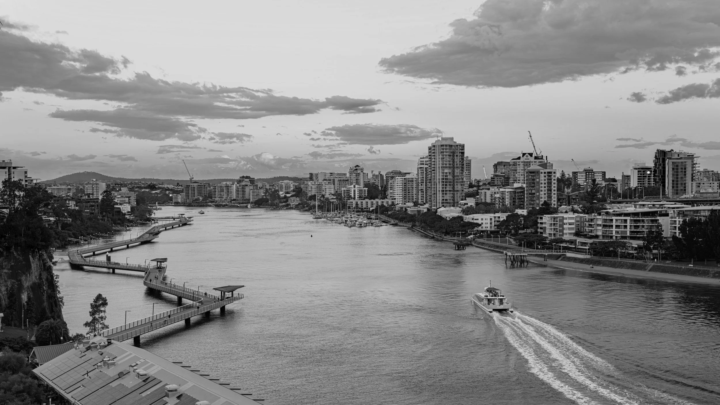 A black-and-white photograph of a city skyline with high-rise buildings along a river, boats on the water, and a sky with scattered clouds.