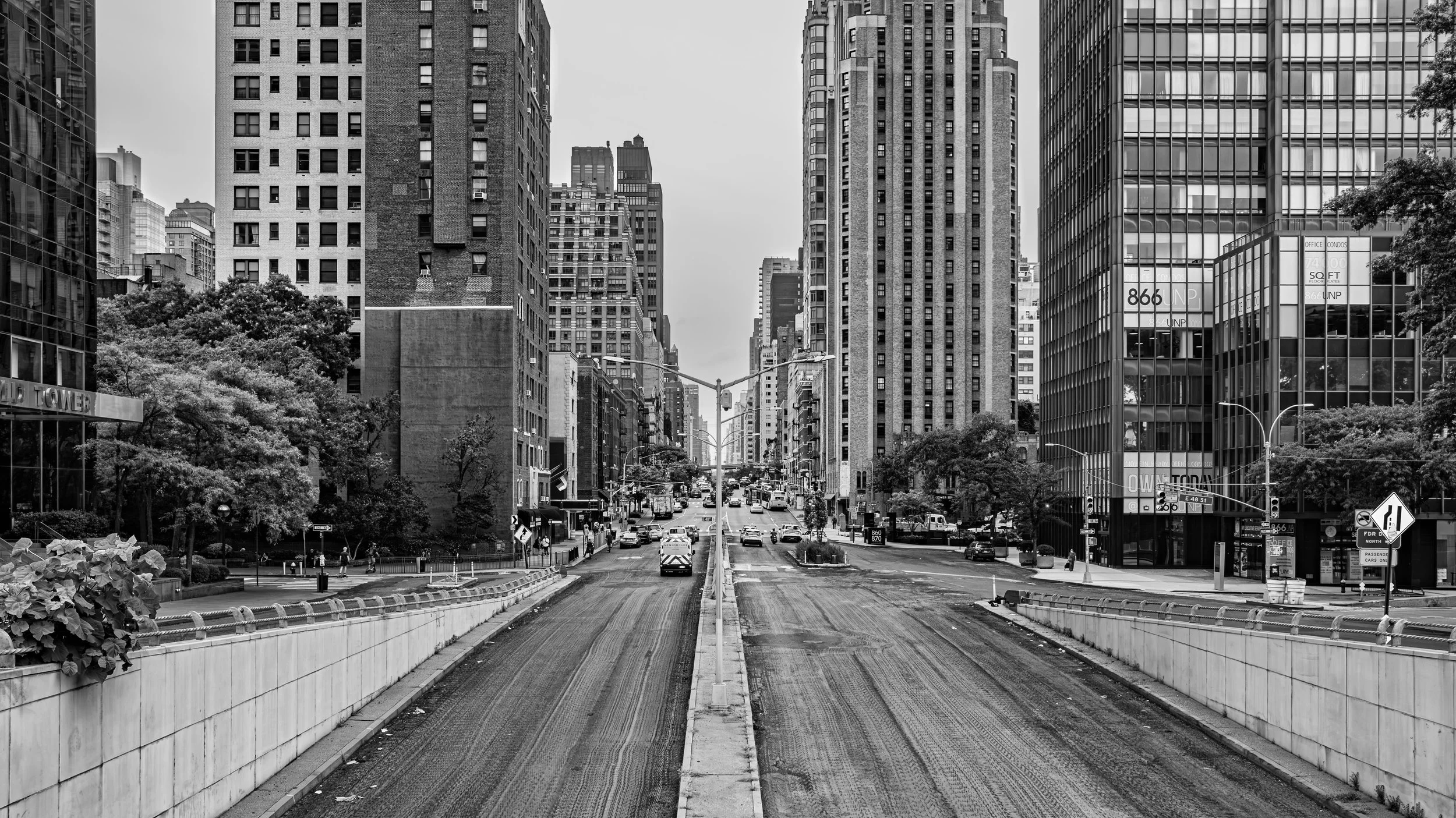 Black and white photo of a city street with tall buildings on both sides, some trees, traffic, and construction barriers and vehicles in the foreground. Manhattan, New York, USA.