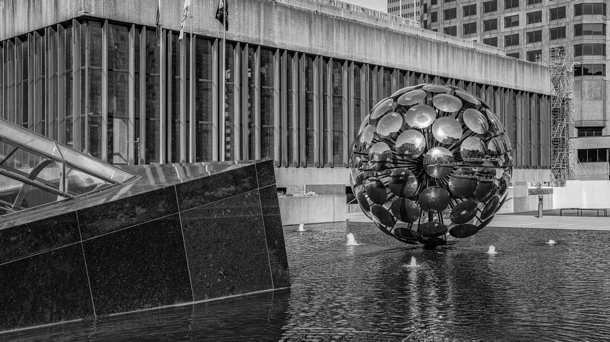 Black-and-white photo of a modern building with tall glass windows and a large reflective spherical sculpture in a shallow water fountain in the foreground.