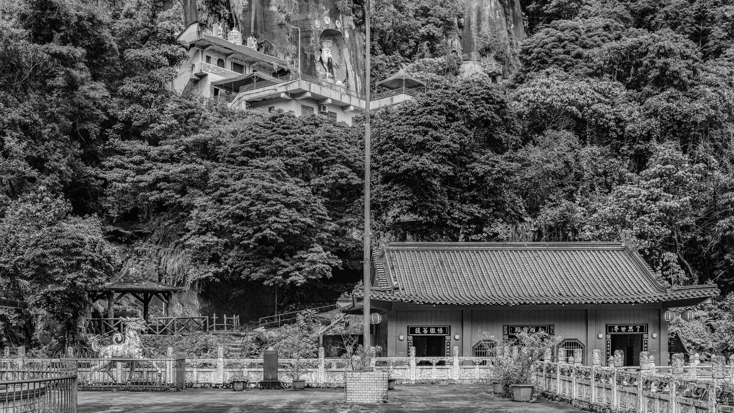 A temple with a sloped tile roof surrounded by decorative fences and potted plants, located in front of a lush, forested hillside with large trees and a statue of a deity or historical figure carved into the rock face.