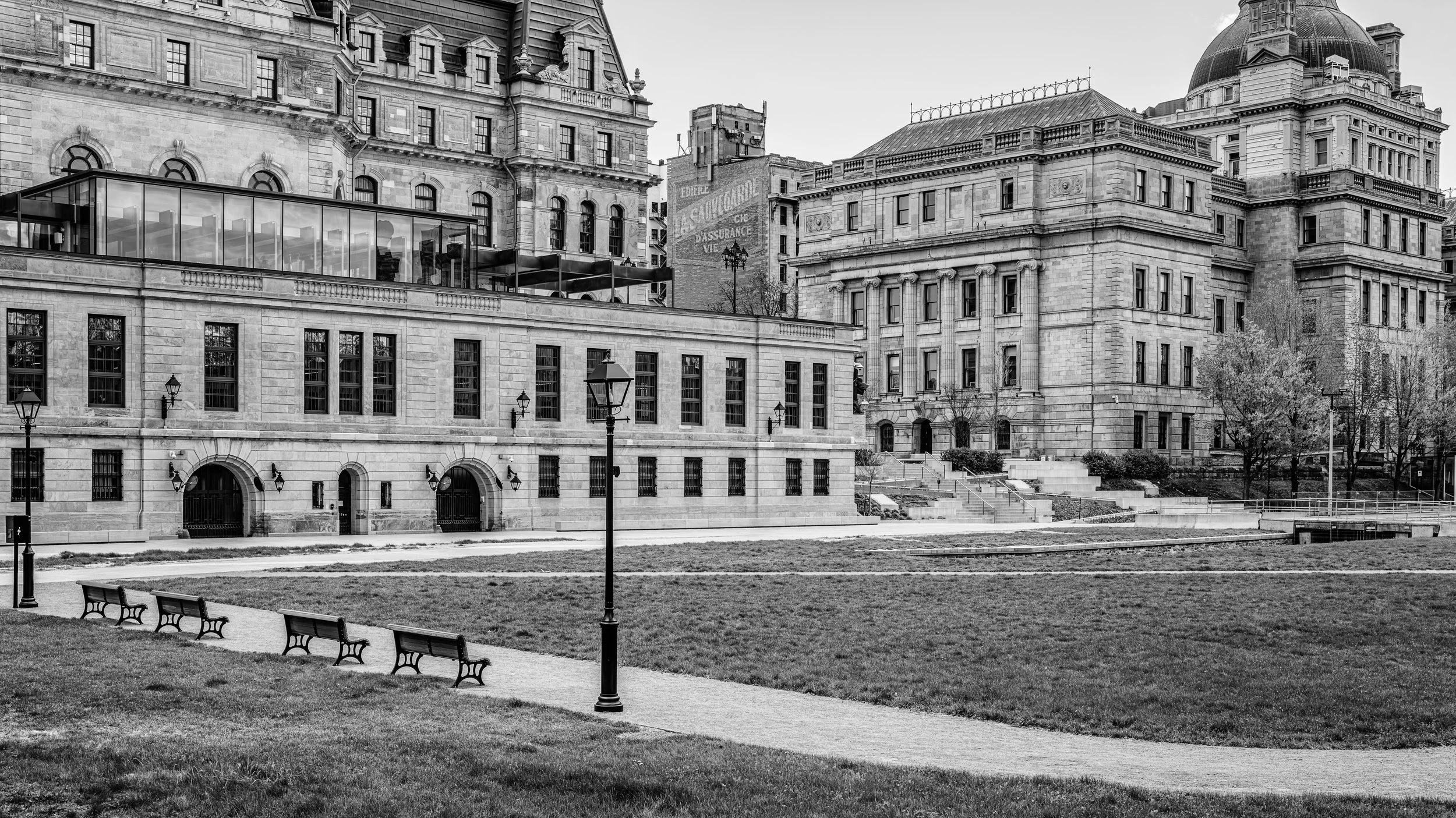 A black and white photo of a city scene with historic buildings, a park with benches, lamp posts, and a grassy area in the foreground.