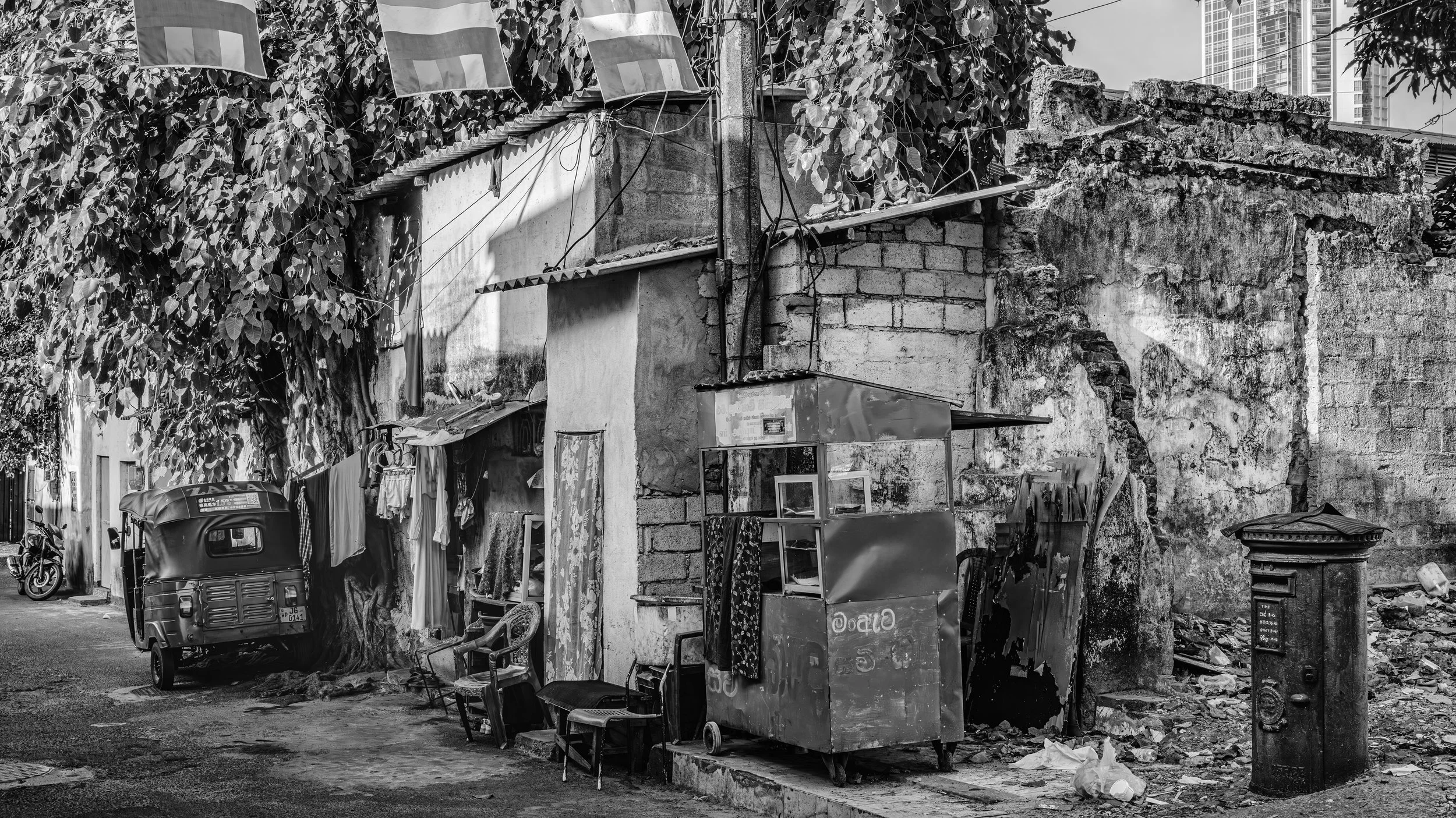 A rundown urban alley with a dilapidated brick wall, an old structure with hanging laundry, a three-wheeled vehicle, a motorcycle, a small food cart, and various debris and trash on the ground.