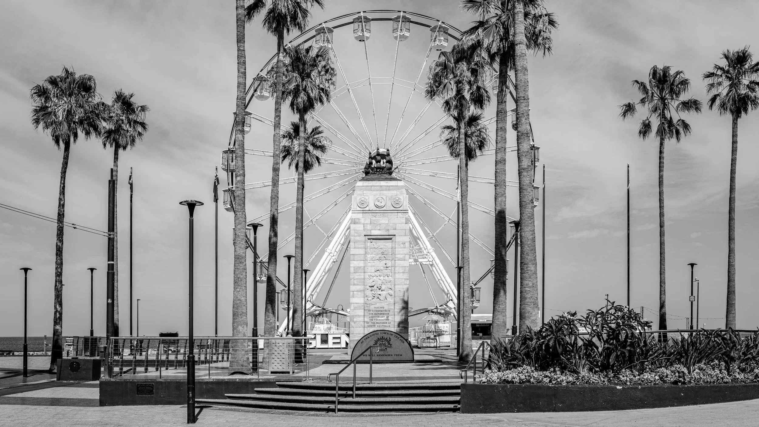 A black-and-white photo of a large Ferris wheel surrounded by tall palm trees, with a monument in front and a partly cloudy sky in the background.